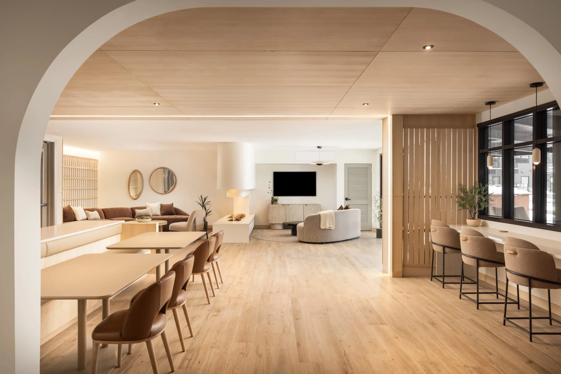 A modern, minimalist lounge with light wood flooring and ceiling. There is a sitting area with sofas, a coffee table, and decorative mirrors on the wall. To the right, there is a counter with bar stools and potted plants near windows. The space has a neutral color palette with warm tones.
