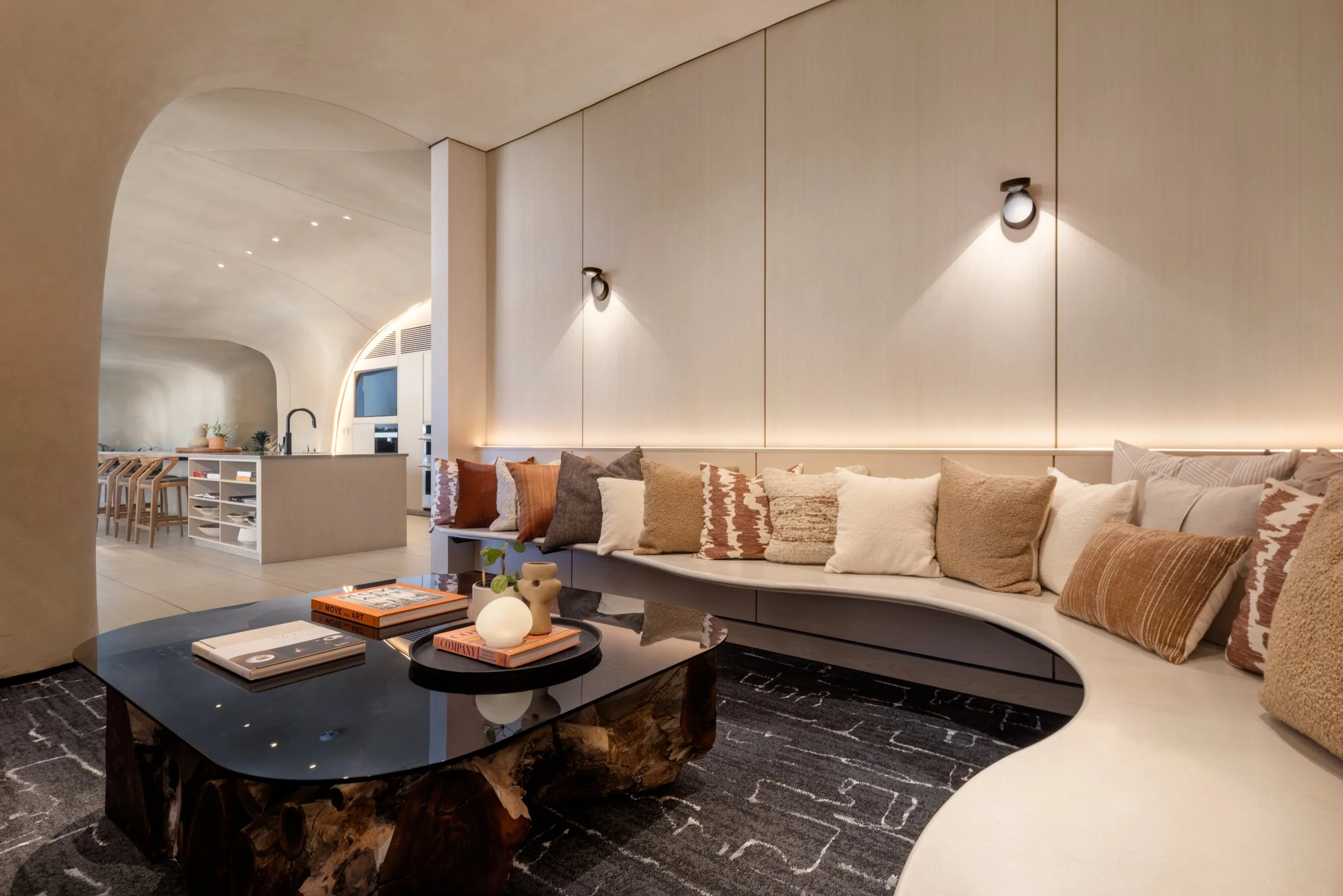 Modern living room with curved ceiling, beige walls, and a long curved sofa with multiple beige and brown pillows. A black glossy coffee table with books and decorative items is in front. The room has soft lighting and an open kitchen area with a white island and chairs.