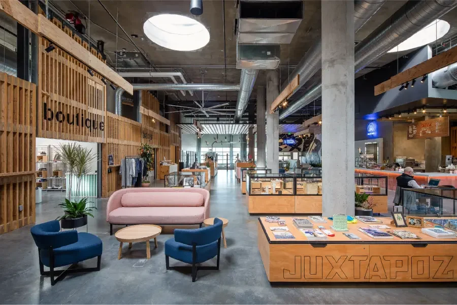 Modern retail store interior with a mix of boutique clothing, jewelry, and magazine displays, a pink sofa with blue chairs and a coffee table in the seating area, industrial decor with exposed ductwork, and a cashier counter.