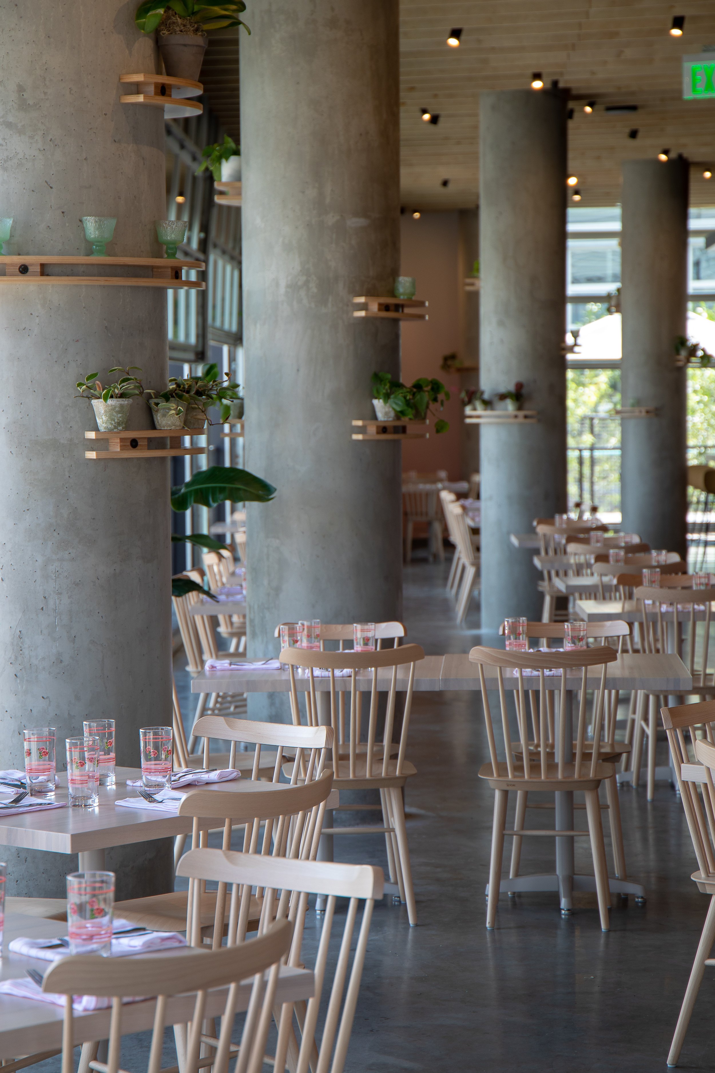 Interior of a modern restaurant with wooden chairs, tables set with glasses and napkins, concrete pillars decorated with potted plants, and large windows letting in natural light.