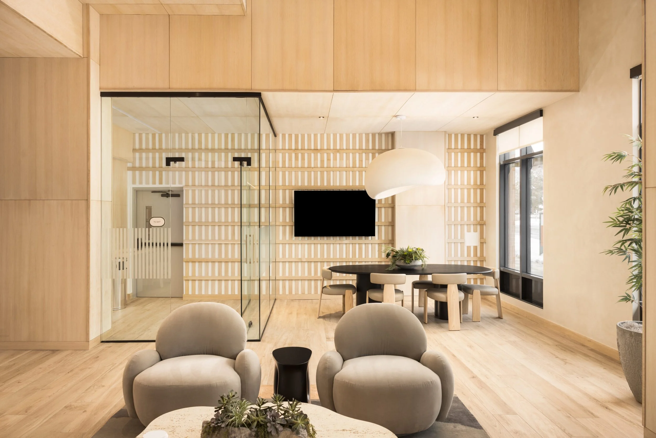 Modern lobby or lounge area with beige armchairs, a black round dining table with beige chairs, a large white pendant light, a flat-screen TV on a wooden slat wall, and large windows with natural light and potted plants.