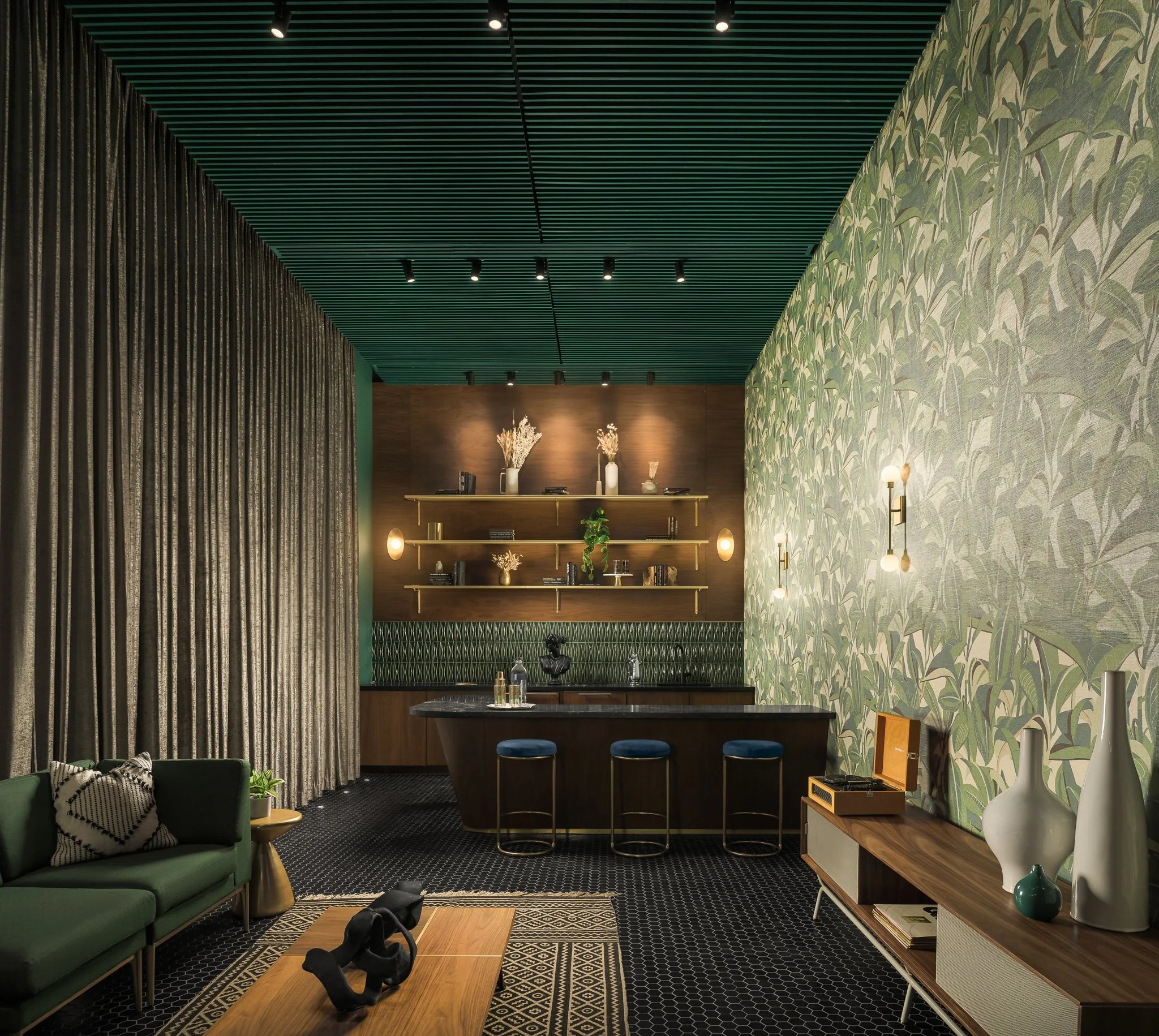 A stylish living room featuring a green sofa with patterned pillows, a wooden coffee table with black abstract sculptures, a textured beige curtain, green patterned wallpaper, a wooden sideboard with decorative vases, and a bar area with blue stools and wall-mounted shelves with decorative items.