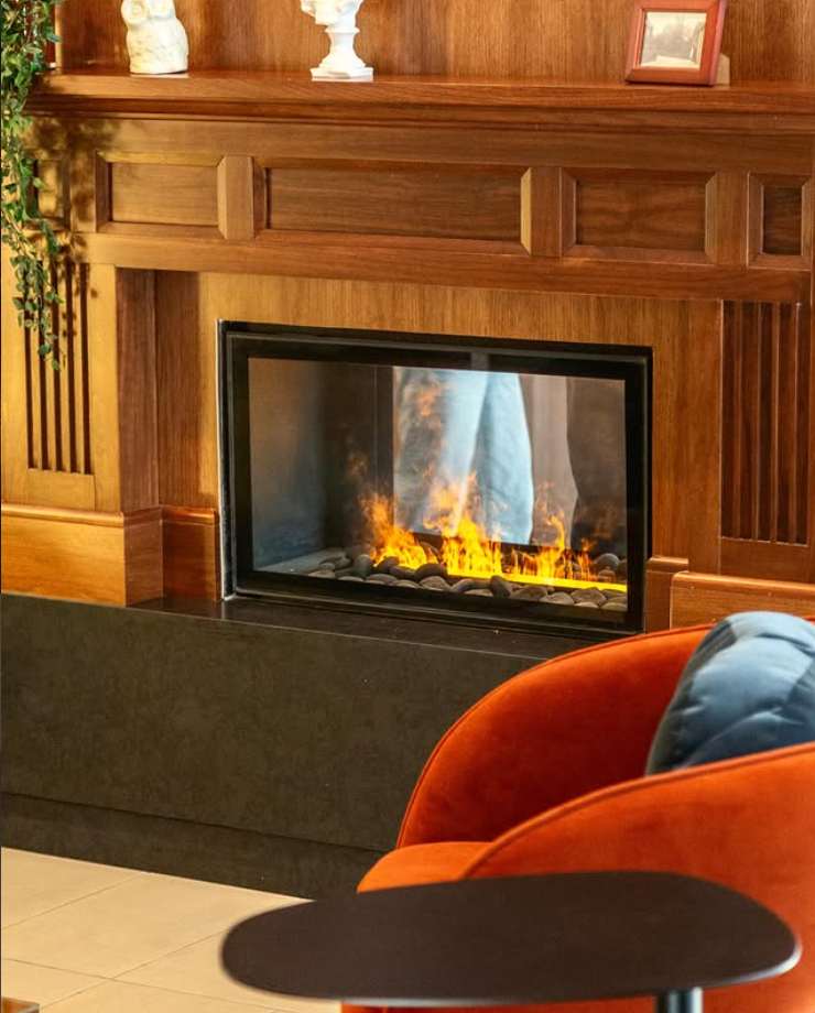 A cozy living room with a wooden fireplace mantel, a glass-enclosed fireplace with flickering flames and black stones, a red armchair with a blue pillow, a small side table, a potted plant, and decorative items on the mantel including a small sculpture and framed photo.