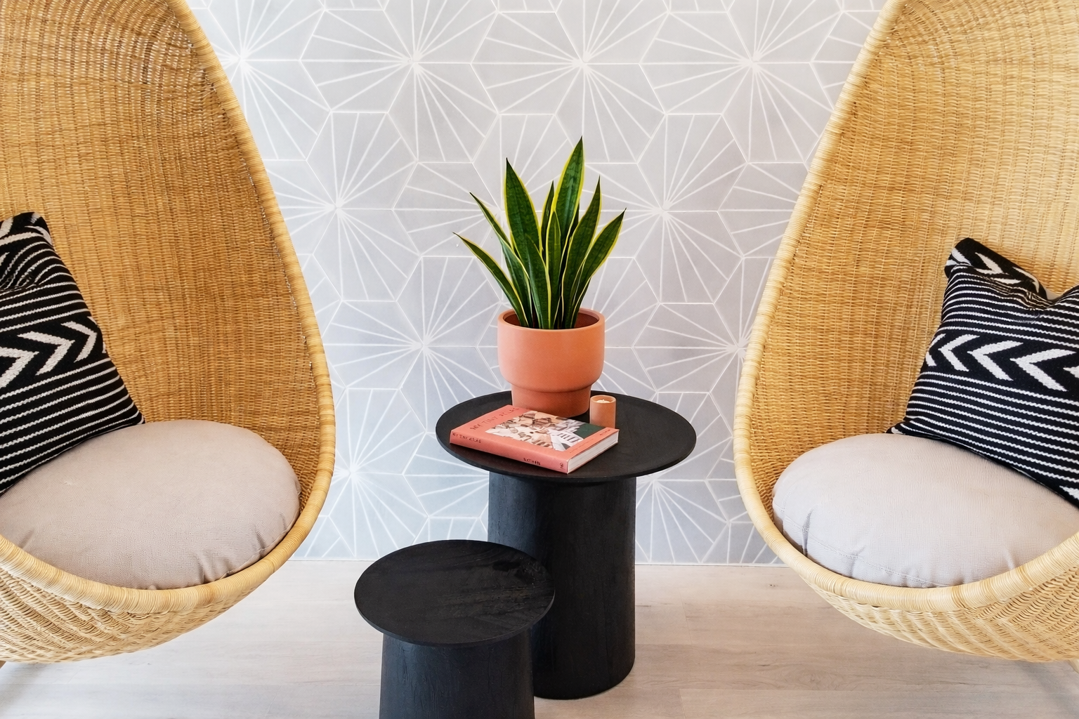 Interior space with two hanging wicker chairs with cushions, a black side table with a potted plant, a book, and a small candle, against a geometric patterned wall.