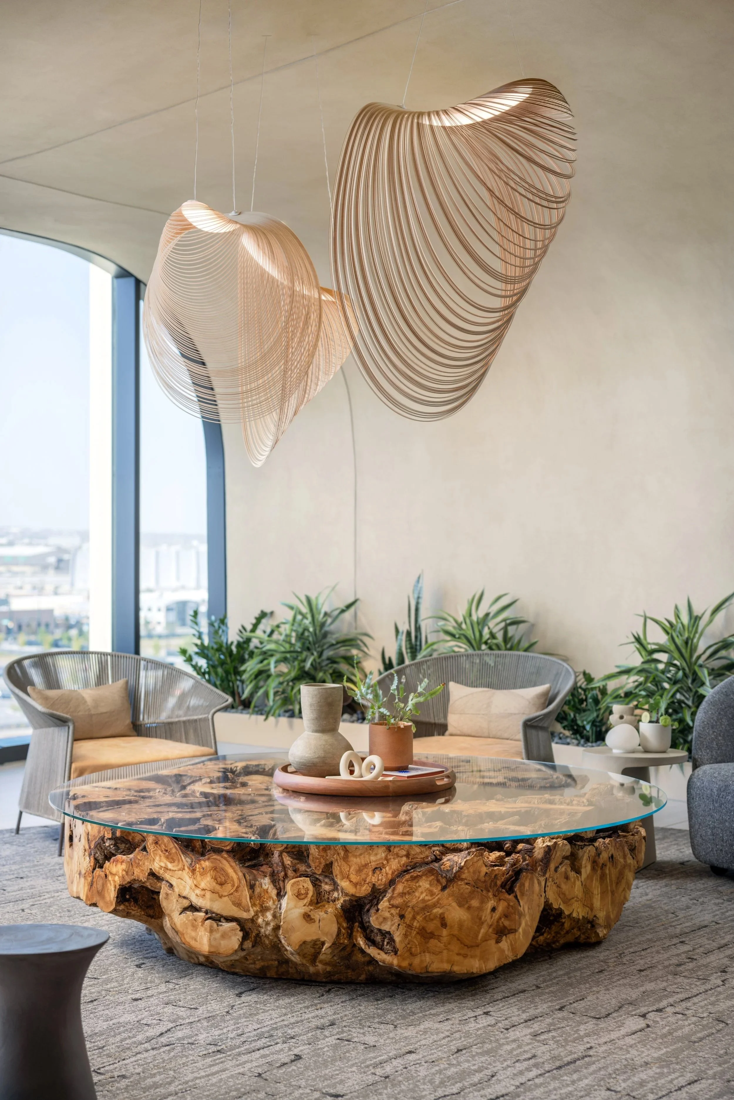 Modern living room with a unique wooden and glass coffee table, two wicker chairs with cushions, potted plants, and large decorative hanging light fixtures.