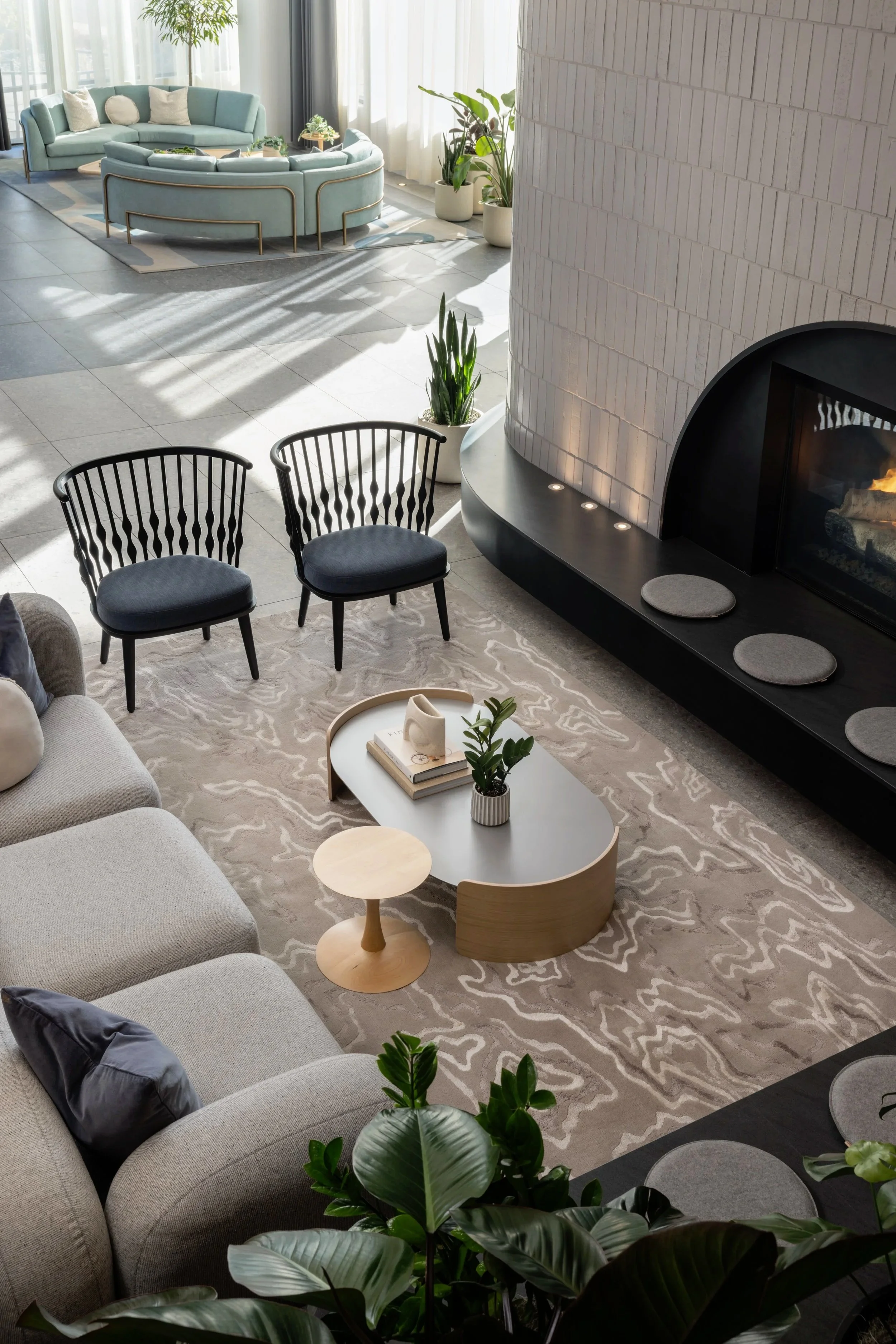 Modern living room with a fireplace, gray sofa, black chairs, a coffee table with books and a plant, and a teal curved sofa in the background near windows with curtains.