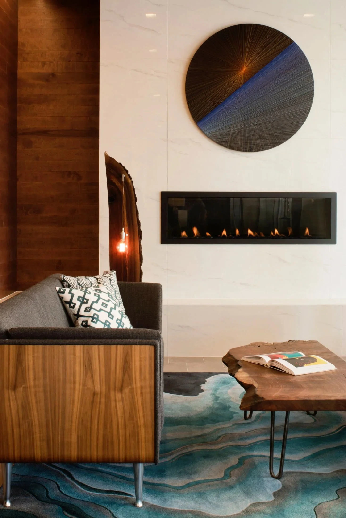 Modern living room with a gray sofa, geometric patterned pillows, a wooden coffee table, an abstract round wall art piece, a linear fireplace, and a textured area rug.