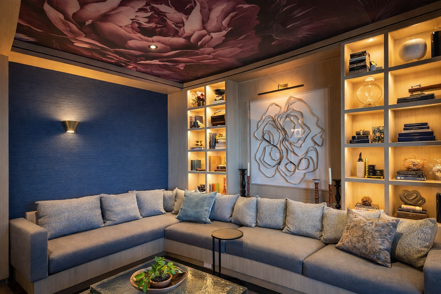 Contemporary living room with a large gray sectional sofa, blue accent wall, built-in wooden shelves with decorative items and books, abstract wall art, floral ceiling mural, and warm lighting.