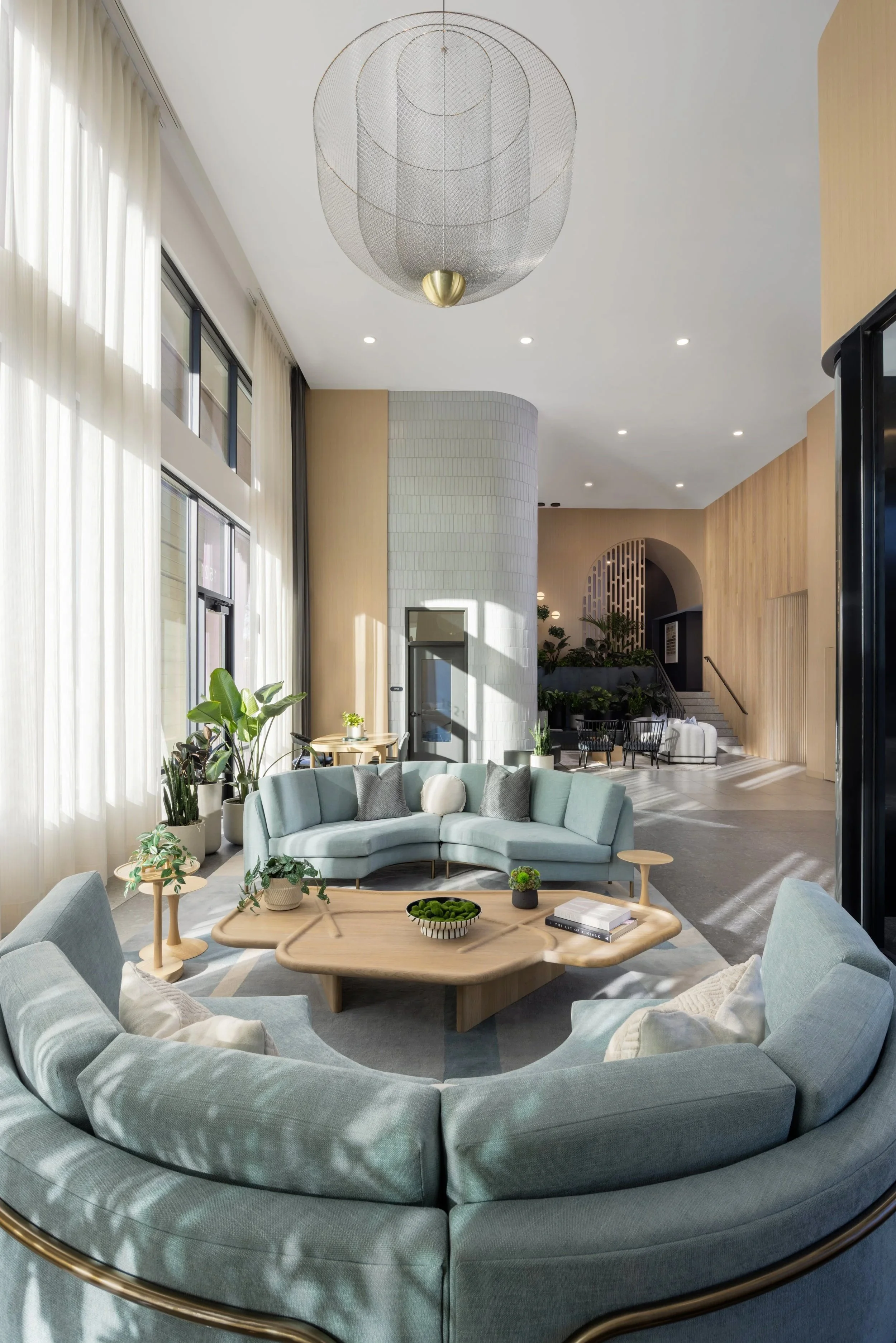Bright, modern living room with a curved light blue sofa, grey armchair, tall windows with white curtains, wooden coffee table with books and plants, and various potted plants near the windows.