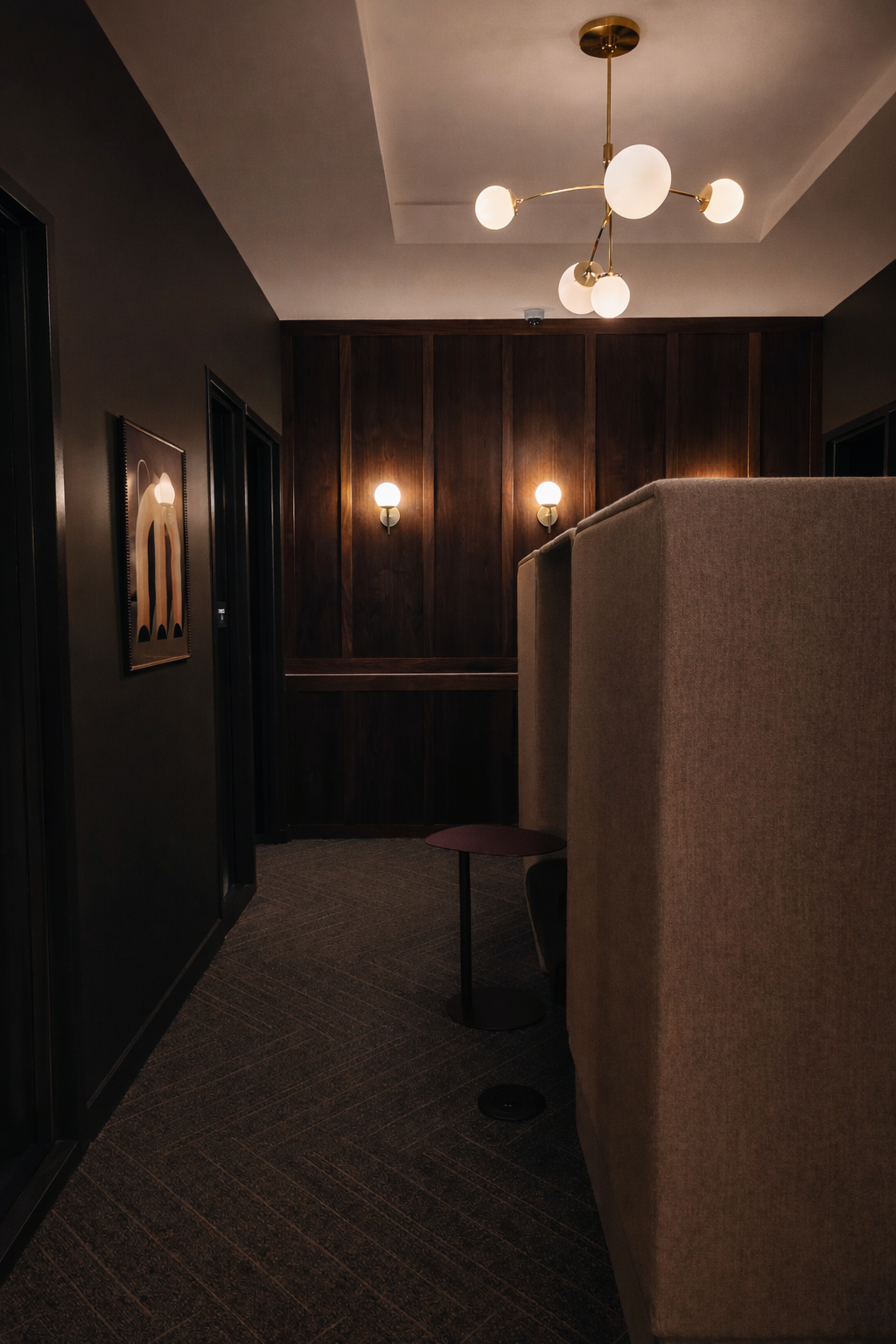 Cozy seating area with beige high-back chairs, dark wood wall paneling, and modern lighting fixtures, including a ceiling chandelier with white globes and wall sconces.