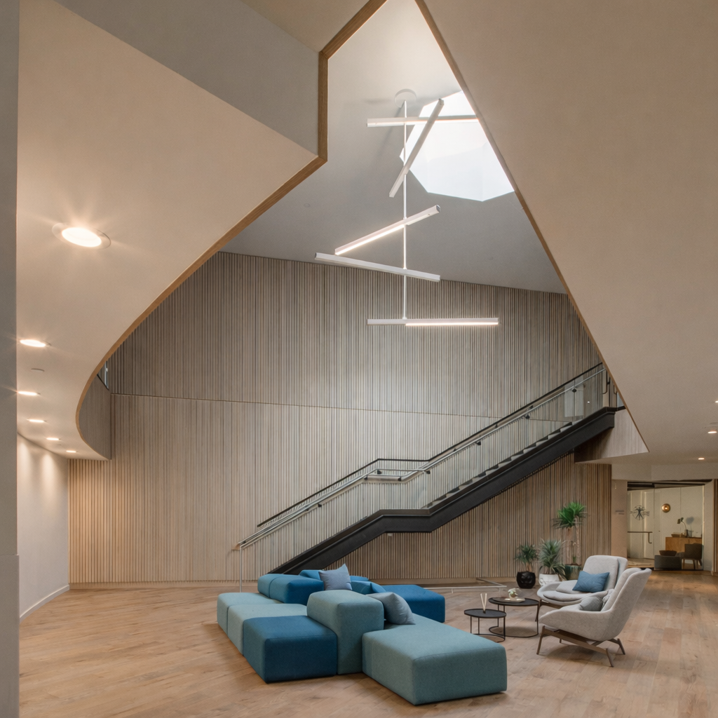 Modern lobby area with a wood-paneled wall, a staircase with glass railings, and a seating area with a blue sectional sofa and two beige armchairs. There are potted plants and contemporary light fixtures hanging from the ceiling.