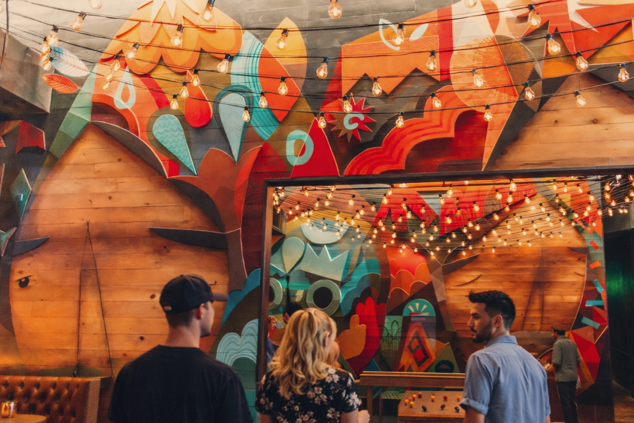 A colorful mural with abstract shapes and patterns and string lights hanging overhead inside a lively indoor setting. Four people are seen conversing in the foreground, with one playing foosball.