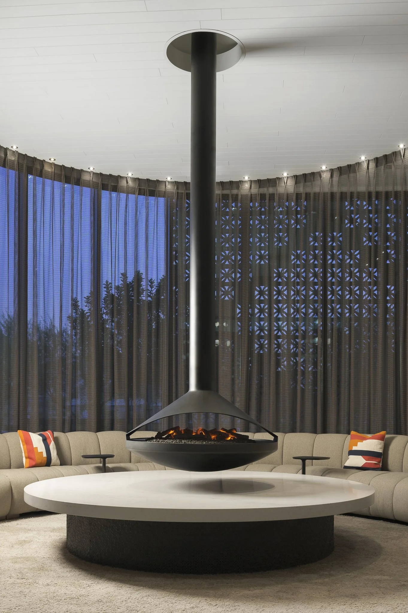 Modern living room with a hanging fireplace centered above a round white table, beige sectional sofa with colorful pillows, and large window with dark curtains.