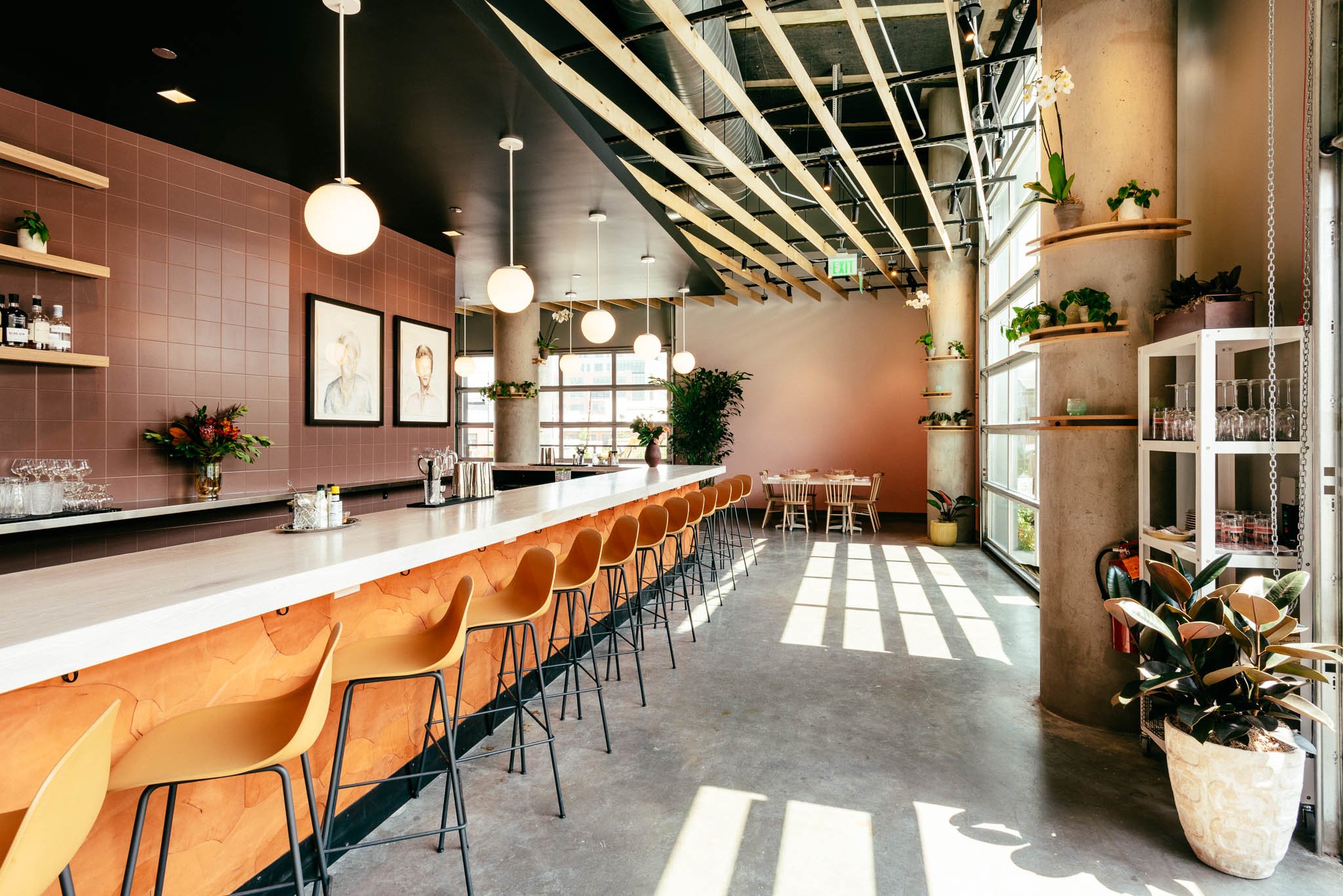 Modern cafe interior with a long bar counter and yellow chairs, decorative plants, large windows allowing sunlight, pendant lighting, and minimalistic decor.