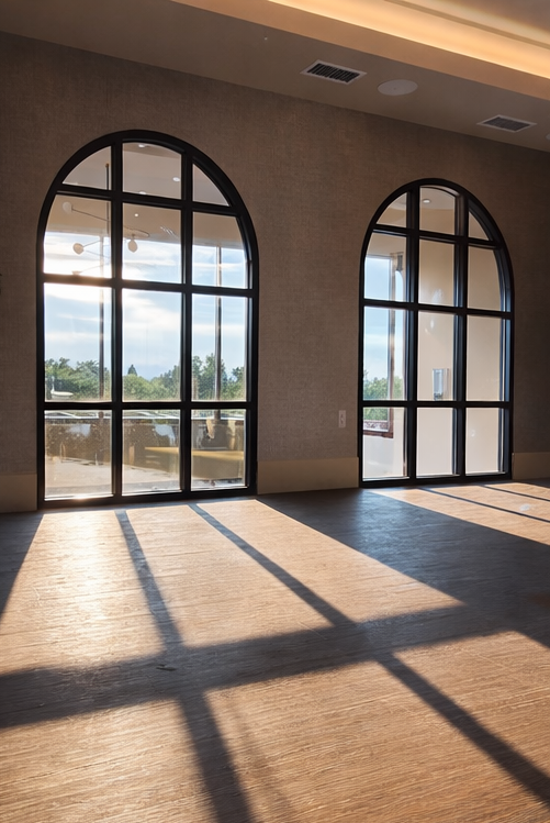 Sunlight streams through large arched windows, casting shadows on the wooden floor inside a modern room with beige walls and ceiling vents.