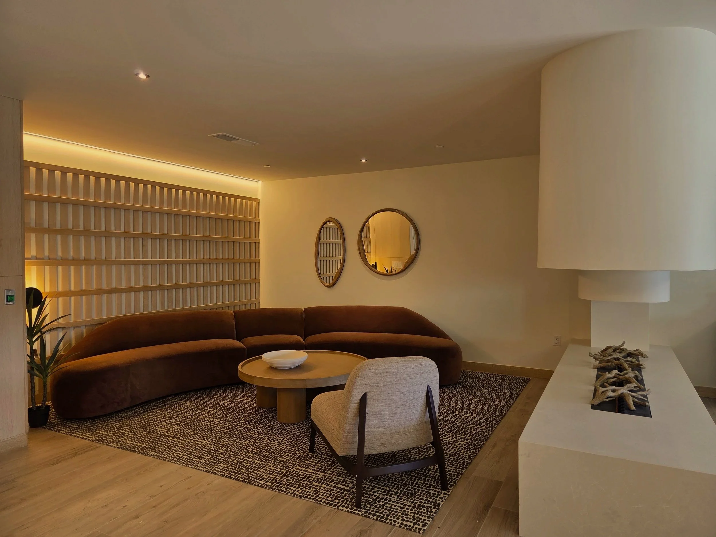 Modern living room with a curved brown sofa, a round wooden coffee table, a beige armchair, and decorative mirrors on the wall.