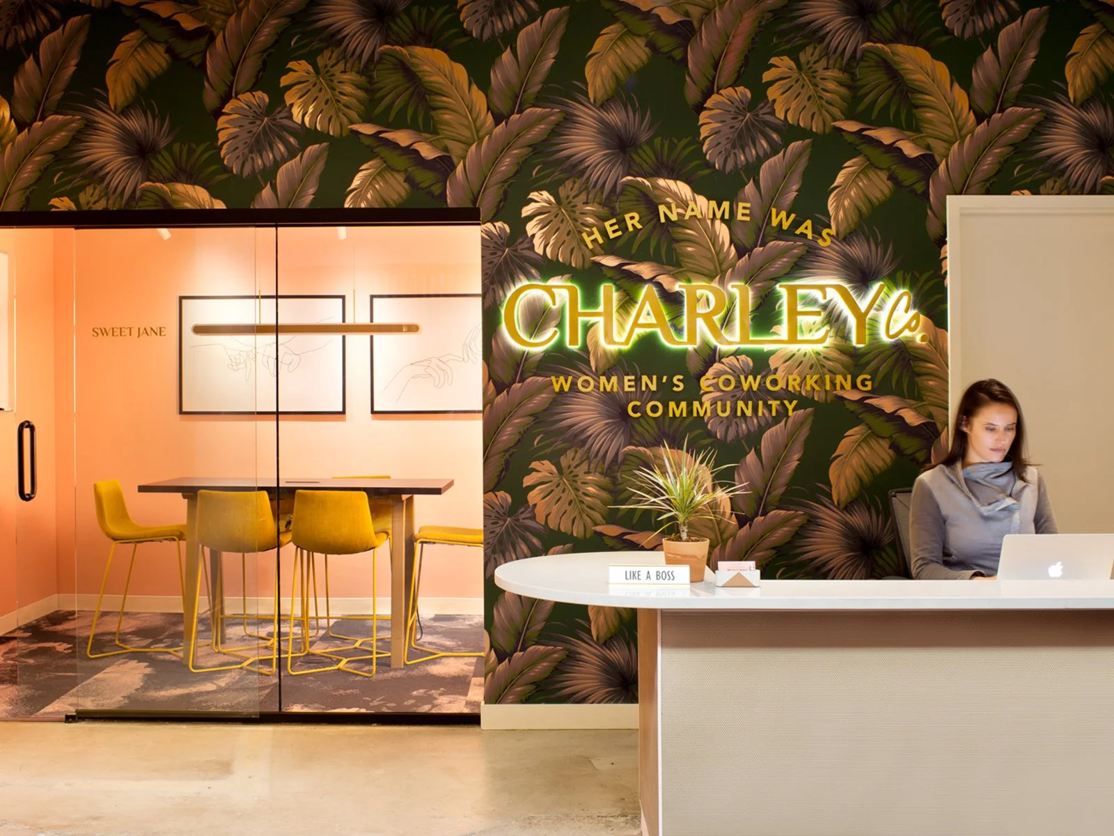 Reception area with a woman working on a laptop behind a white counter, a colorful wall with tropical leaf wallpaper, neon sign reading 'Charley Co.', and a glass door leading to a room with a pink wall, yellow chairs, and a table.
