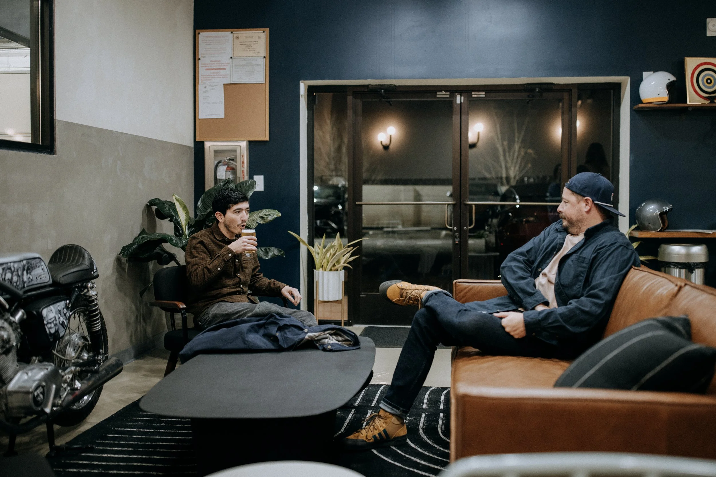 Two men sitting in a cozy lounge, one drinking from a cup with plants and motorcycle nearby, door leading outside in the background.