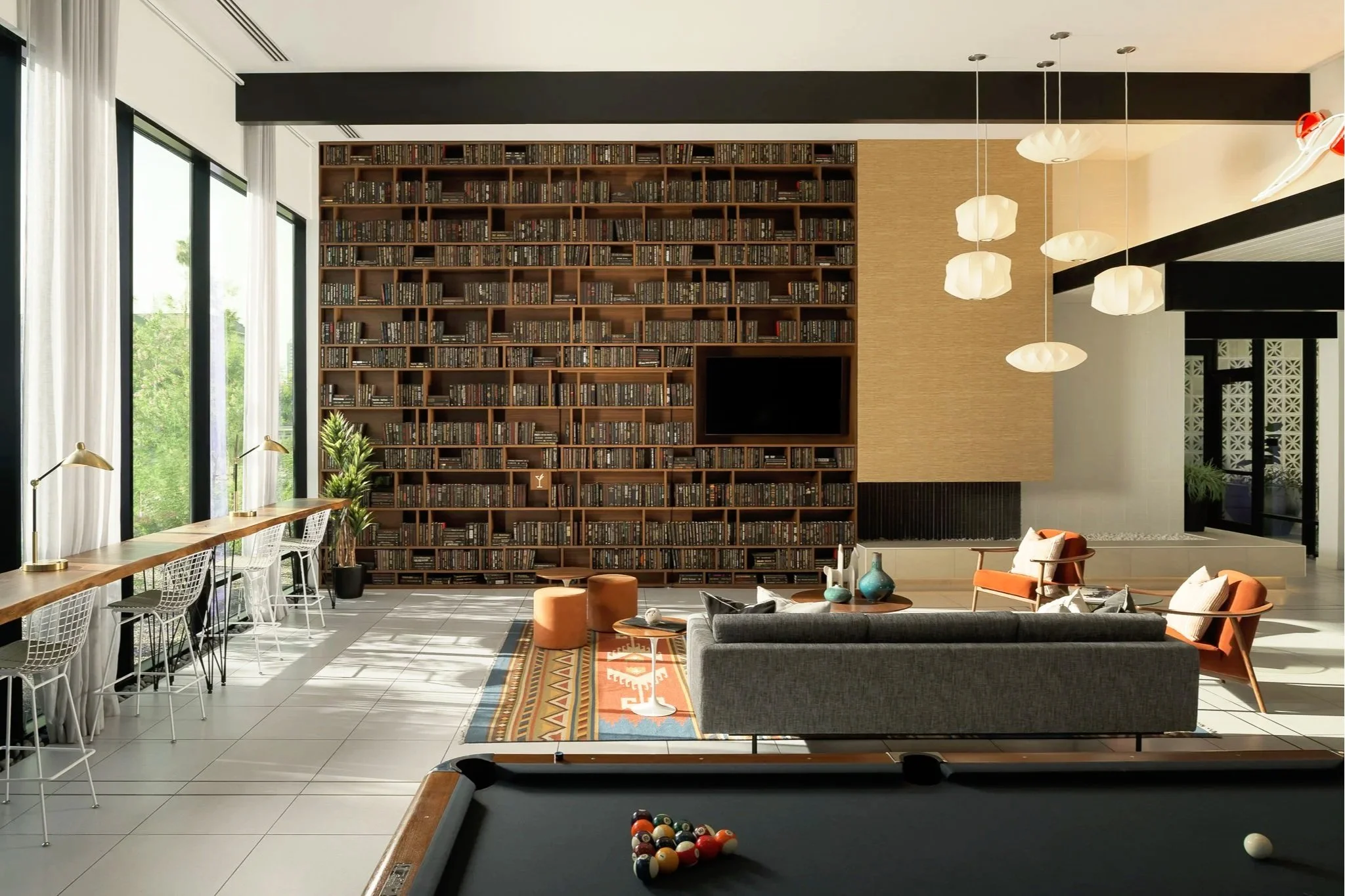 Modern living room with a large wooden bookshelf, sitting area with chairs and a sofa, pool table in the foreground, large windows with white curtains, pendant lights, and a TV mounted on the bookshelf.