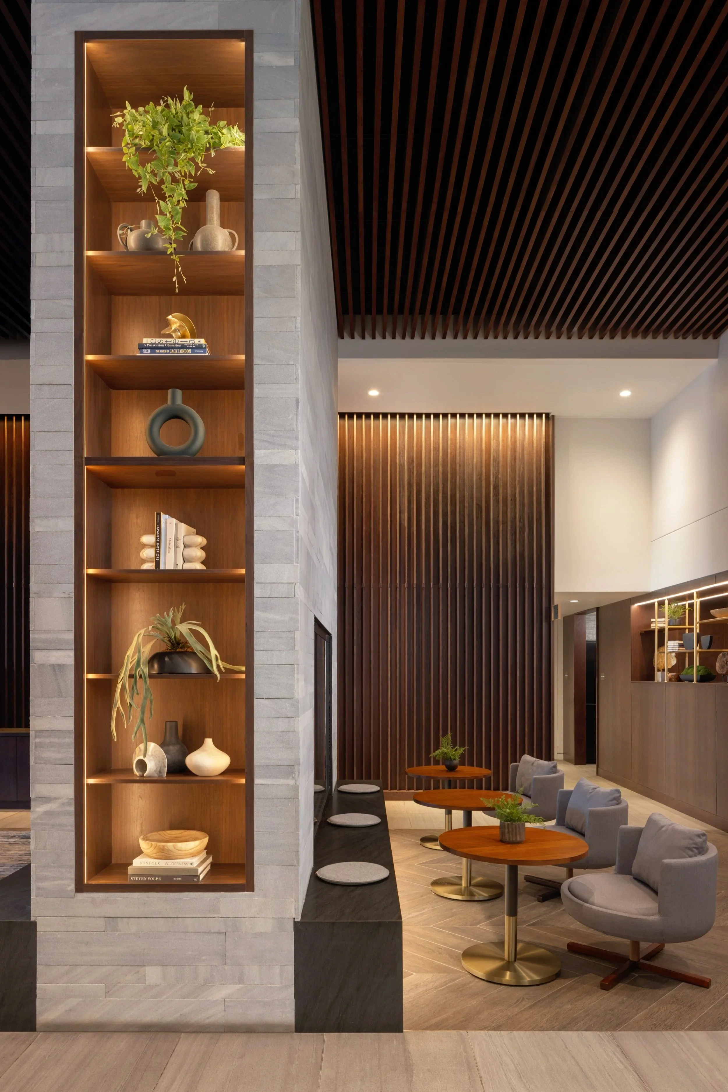 Interior view of a modern lounge with a tall wooden shelving unit displaying decorative vases, books, and plants, adjacent to a seating area with gray armchairs and small round tables.