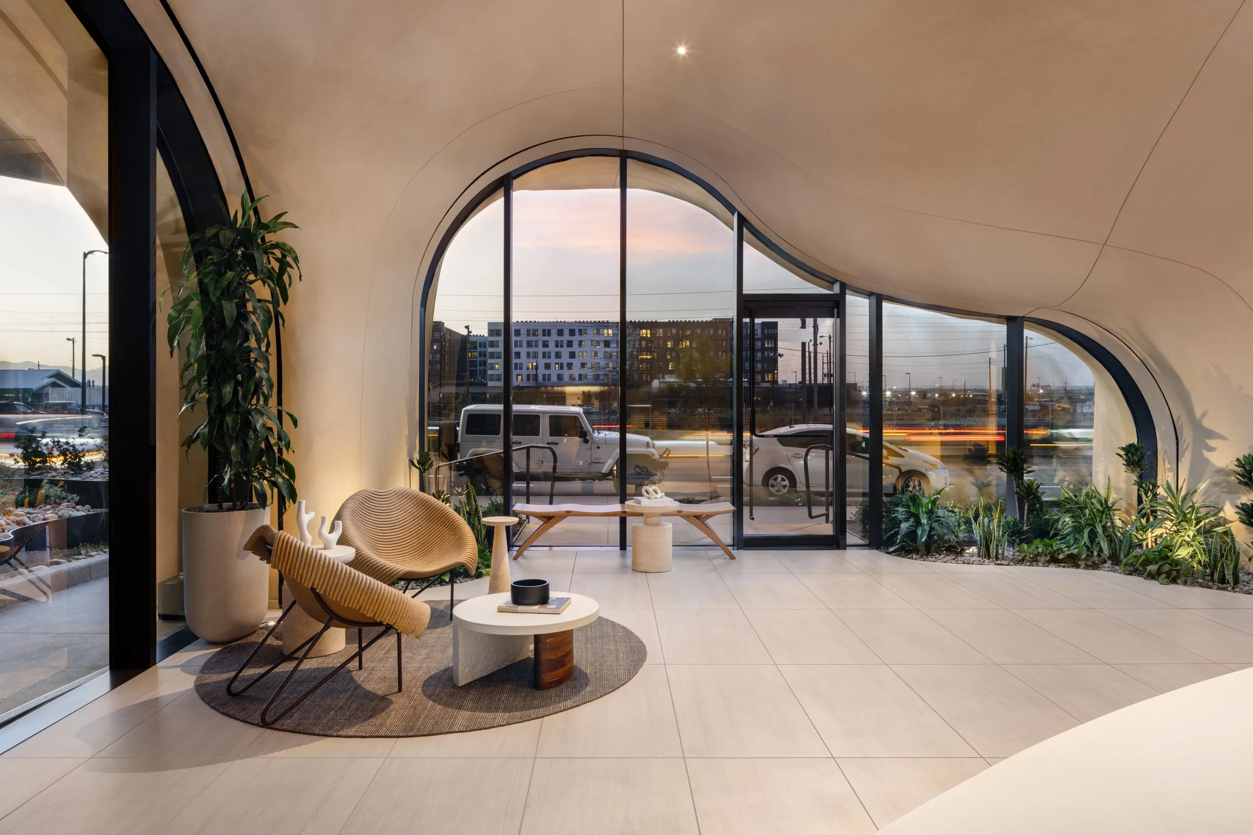 Modern interior with curved ceiling, large glass windows with view of street and moving cars, potted plants, and contemporary furniture including chairs and a coffee table.