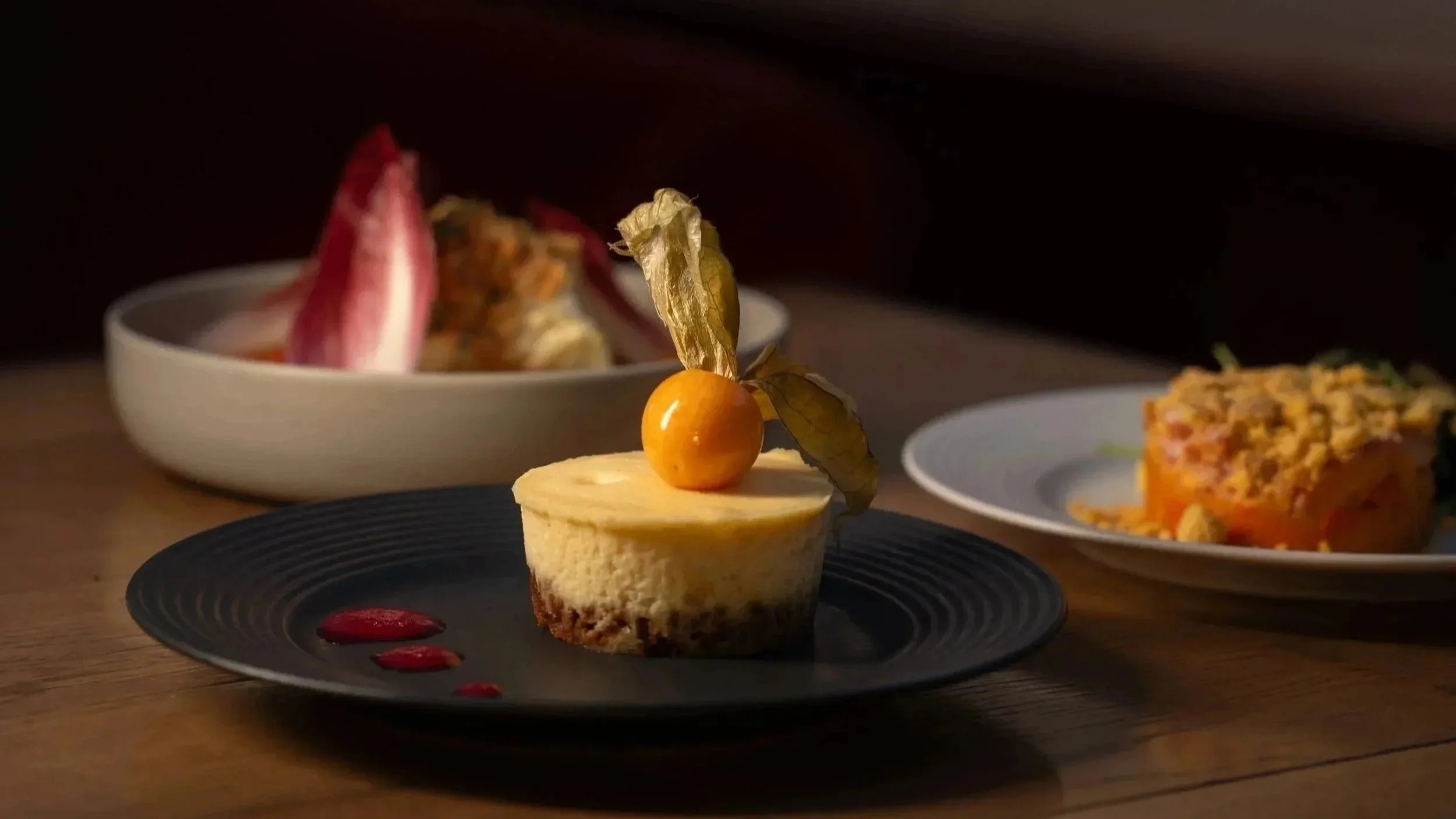 Gâteau au fromage décoré d'une physalis, avec des sauces rouges sur une assiette noire, sur une table en bois, avec deux autres plats en arrière-plan.