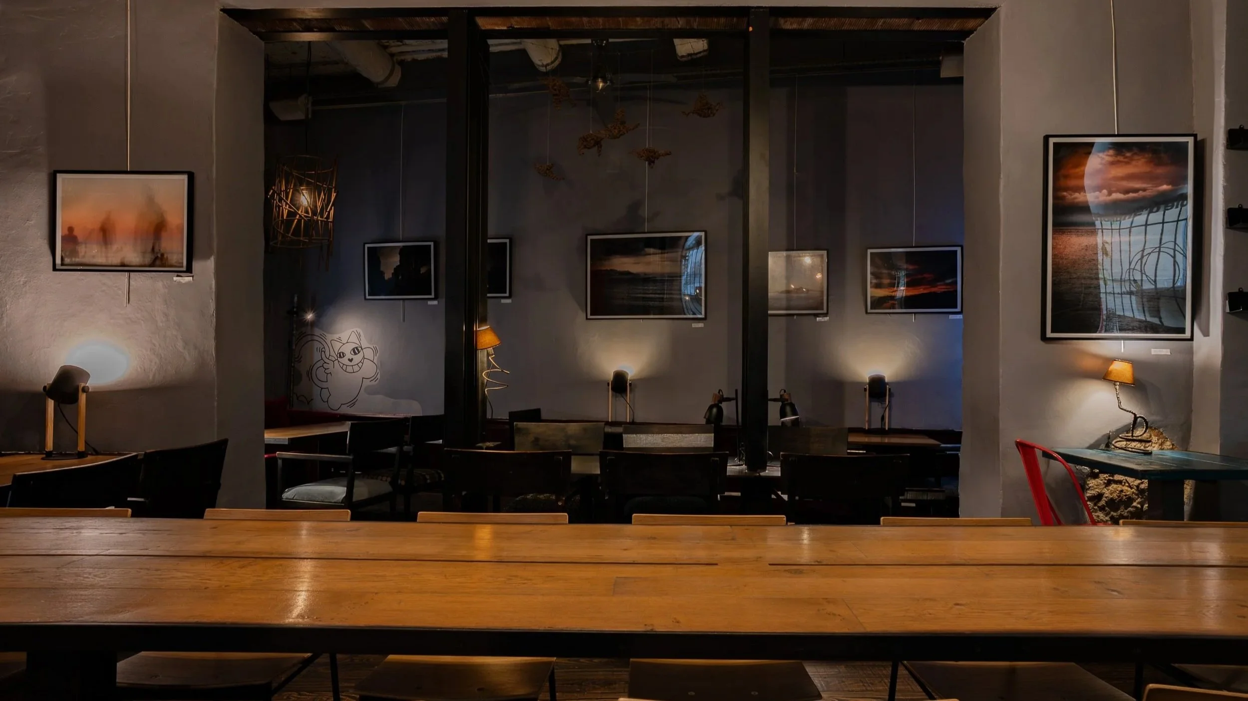 Intérieur d'un café ou d'une galerie d'art avec des tableaux accrochés sur les murs, des lampes d'ambiance, et plusieurs chaises et tables en bois.