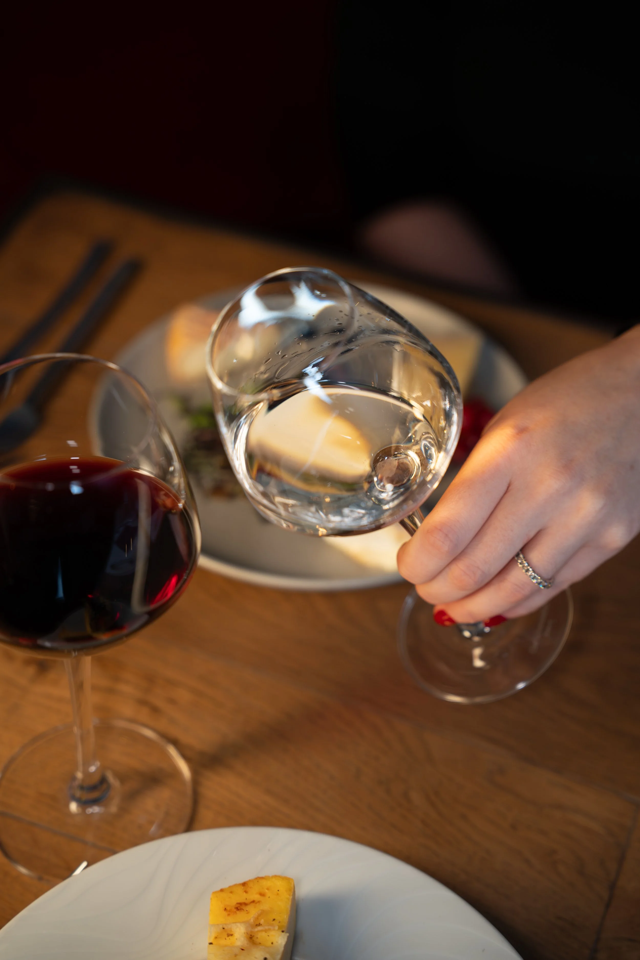 Une personne tenant un verre d'eau, avec verre de vin rouge et un morceau de gâteau sur une table en bois, assiette blanche, fourchette et couteau.