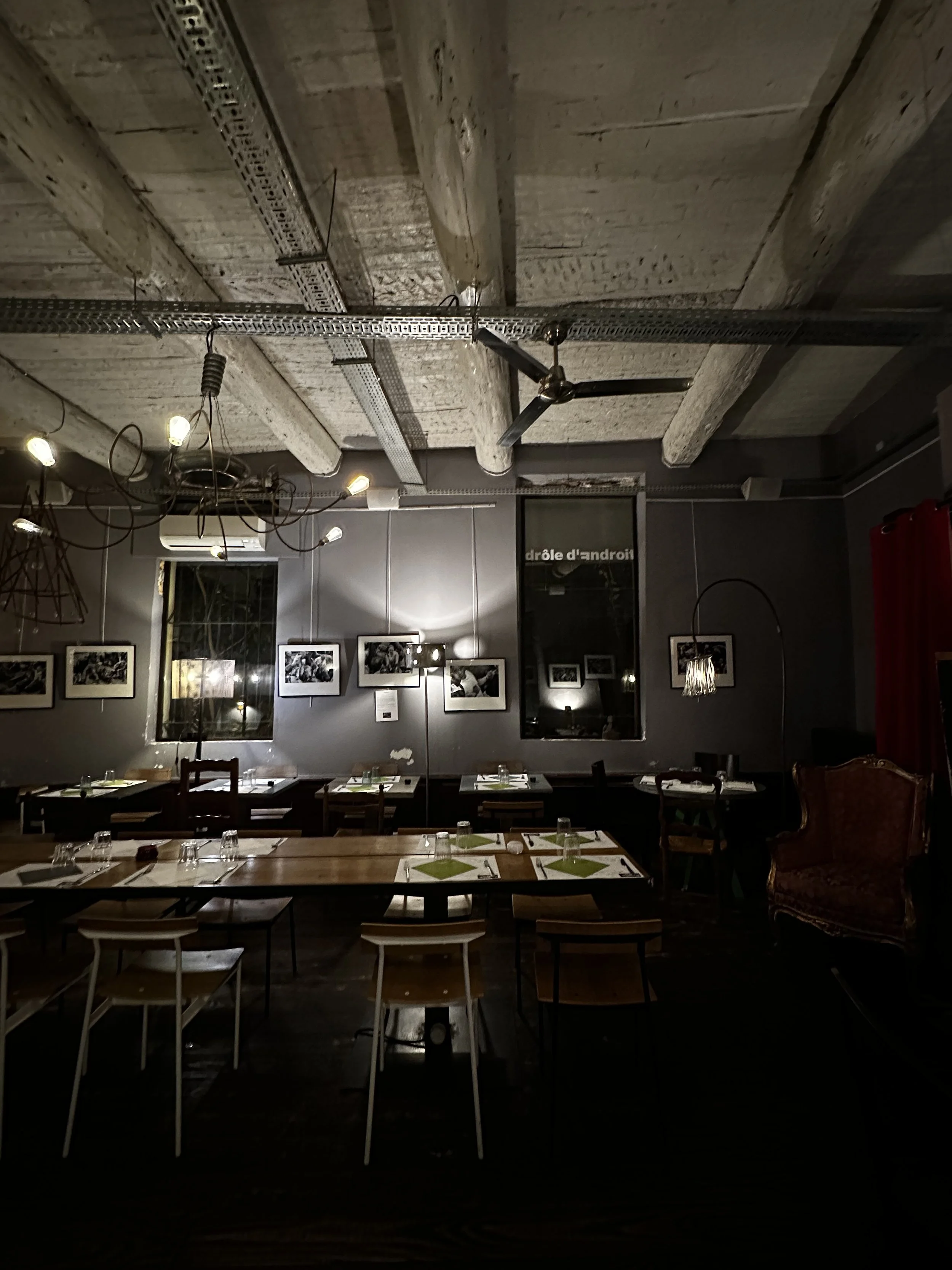 Salle à manger dans un restaurant avec tables en bois, chaises, tableaux photo sur le mur, luminaires et plafond en béton apparent.