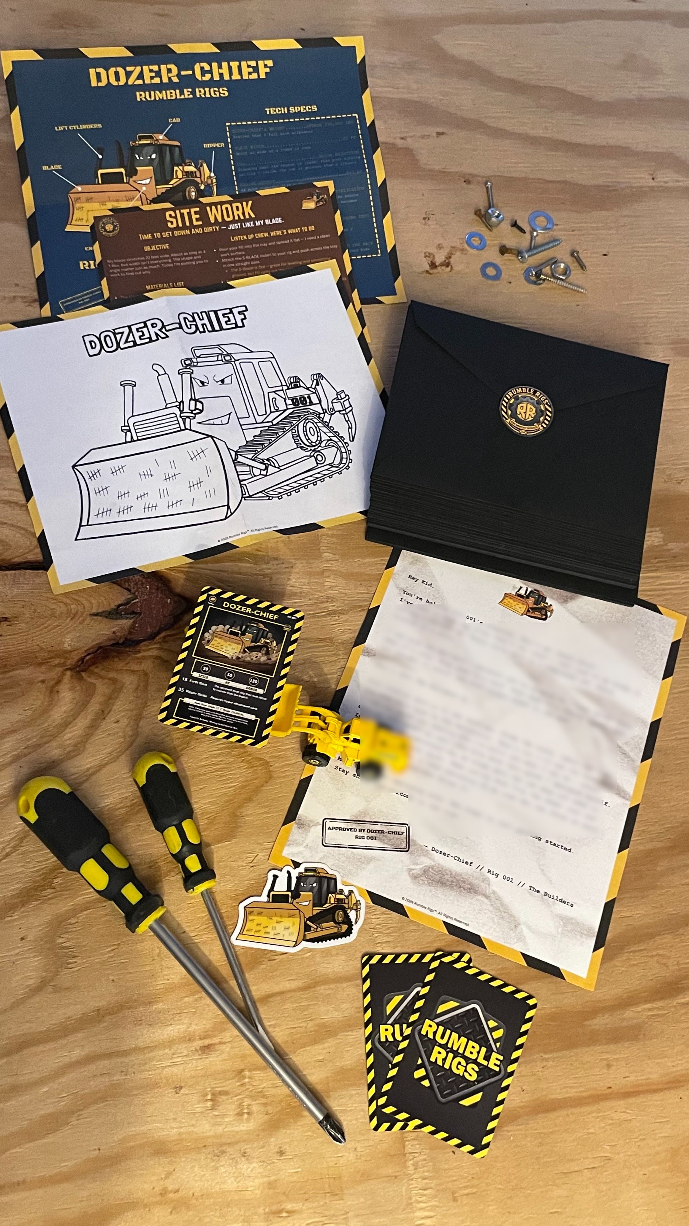 Rumble Rigs monthly dispatch contents — Dozer-Chief trading card, blueprint schematic, coloring sheet, rig letter, die-cut sticker, and black envelope spread out on a workbench.