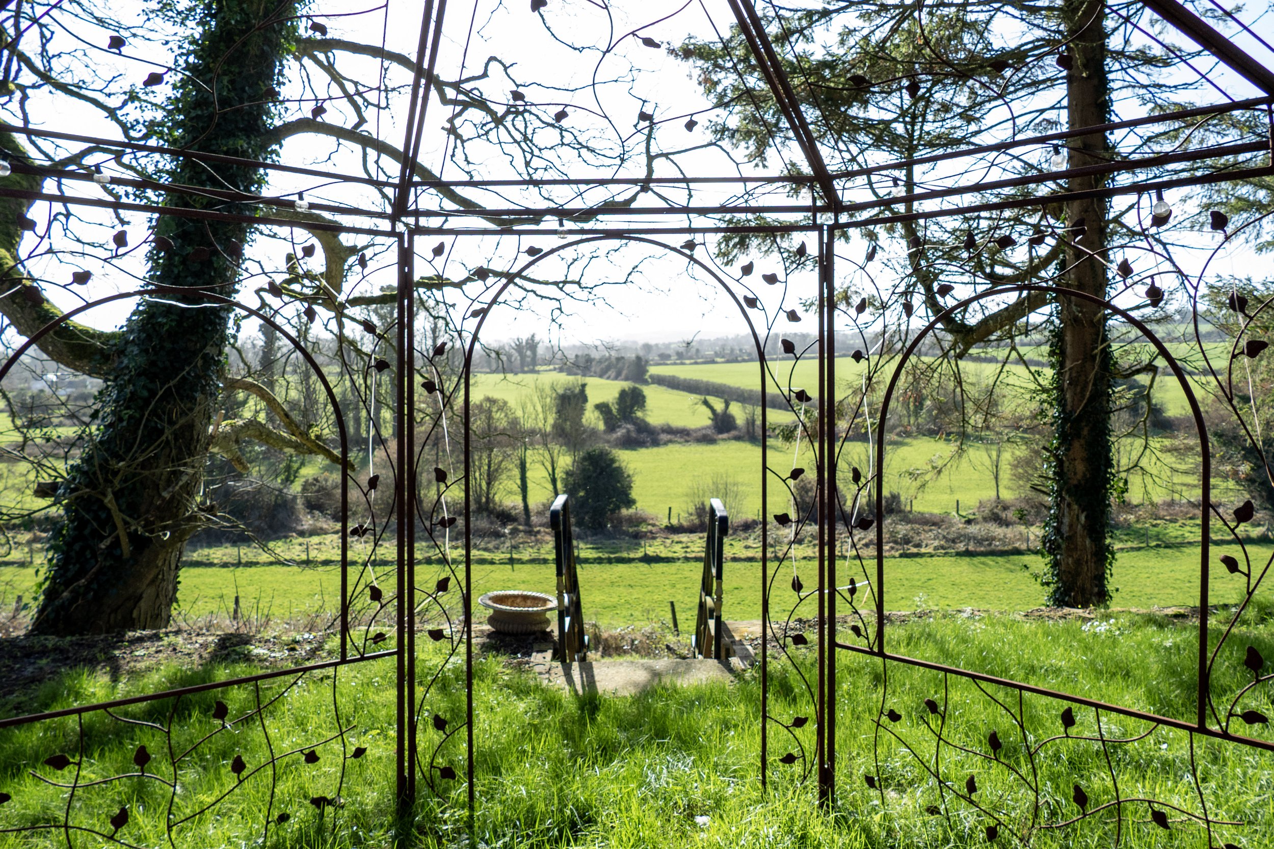 View of a green landscape through a decorative metal arbor with leaf patterns, with stairs and a basket in the foreground.