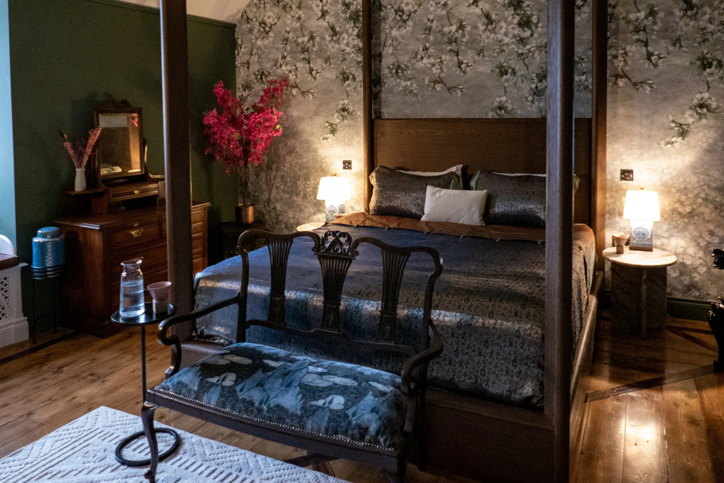 A cozy bedroom with a four-poster bed, floral wallpaper, and bedside lamps. There's a floral-patterned bench at the foot of the bed, a wooden dresser with a mirror, and a small table with water pitcher and glasses. A pink flowering plant is on a side