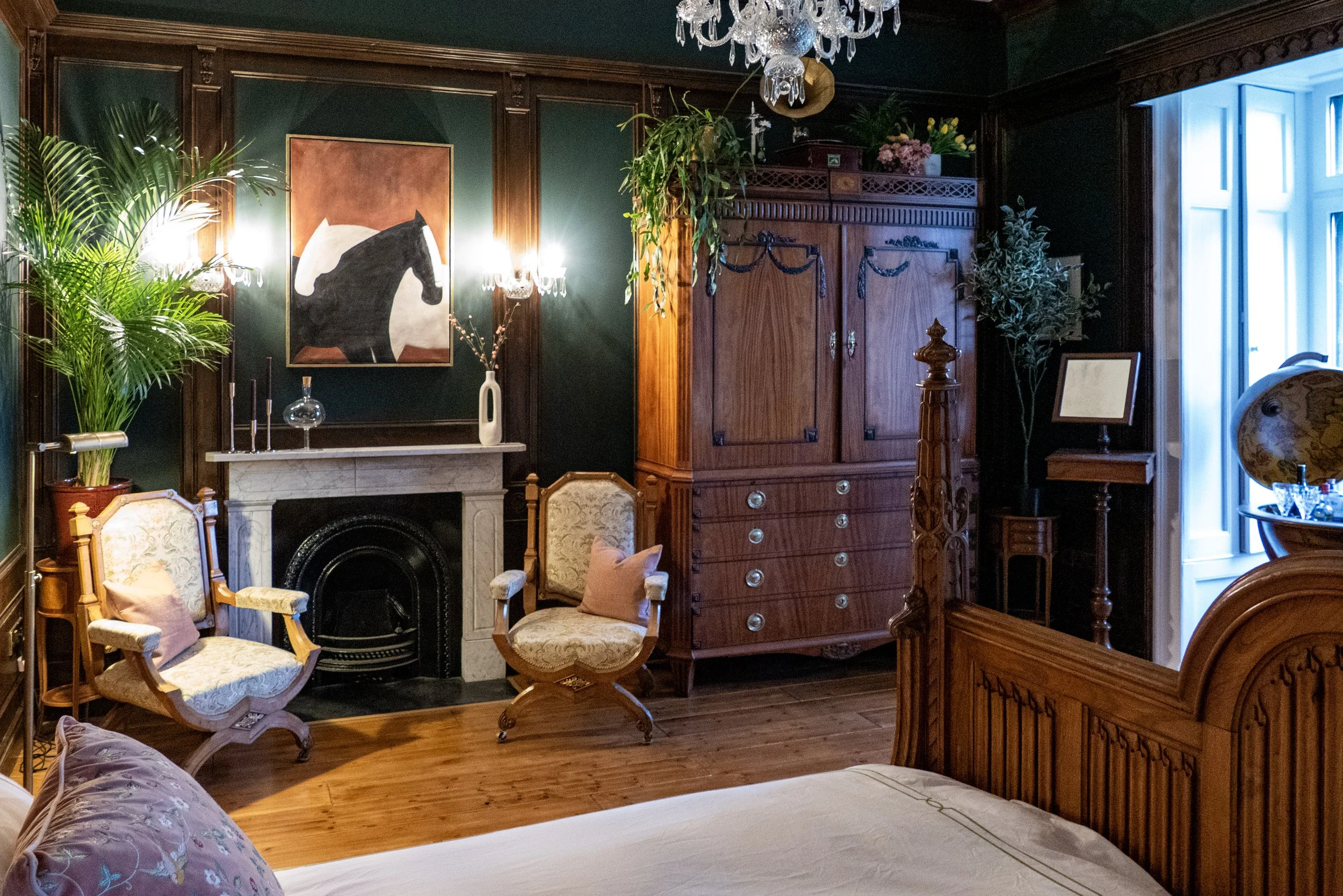 A luxurious bedroom with dark green walls, wooden furniture, a fireplace, two vintage armchairs with pink cushions, potted plants, a large painting of a black horse, and a fireplace mantel decorated with vases and candles.