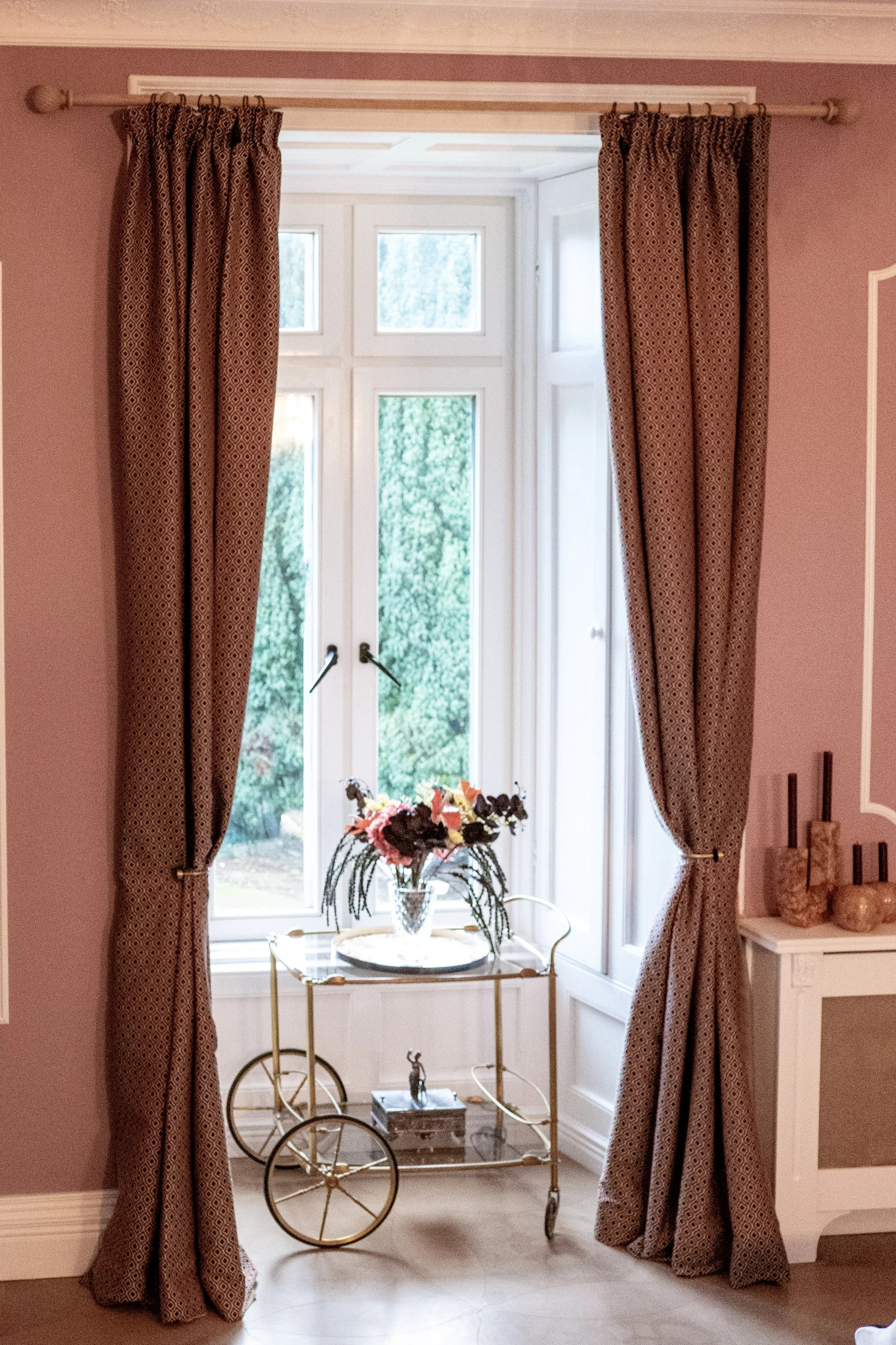 A window with brown patterned curtains, a decorative bar cart with a flower arrangement in a vase, and ceramic decor on a white cabinet in a pink-walled room.