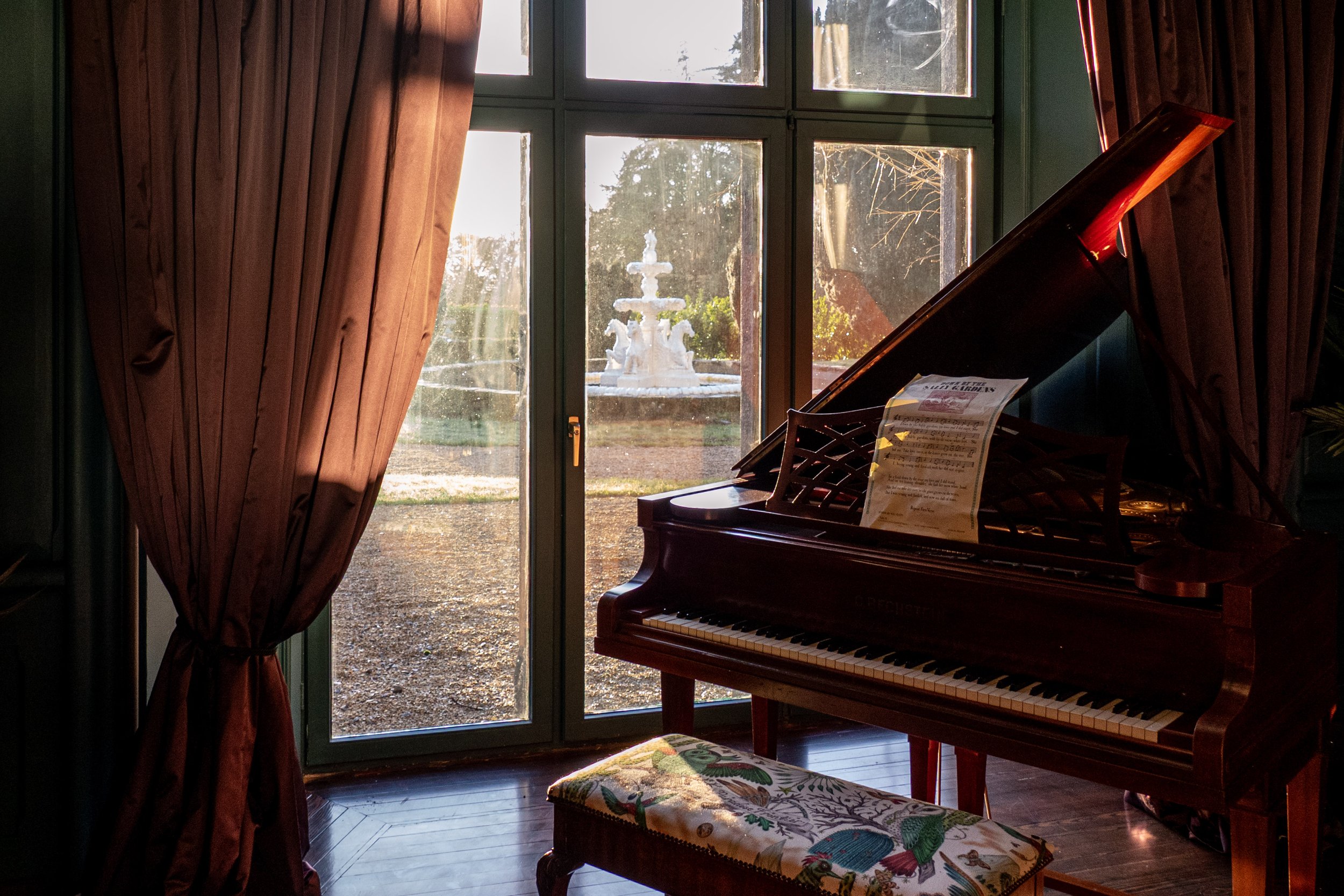 Sunlight streaming through large window with red curtains, highlighting a grand piano with sheet music, and a bench with colorful embroidery, in a room overlooking a garden fountain.