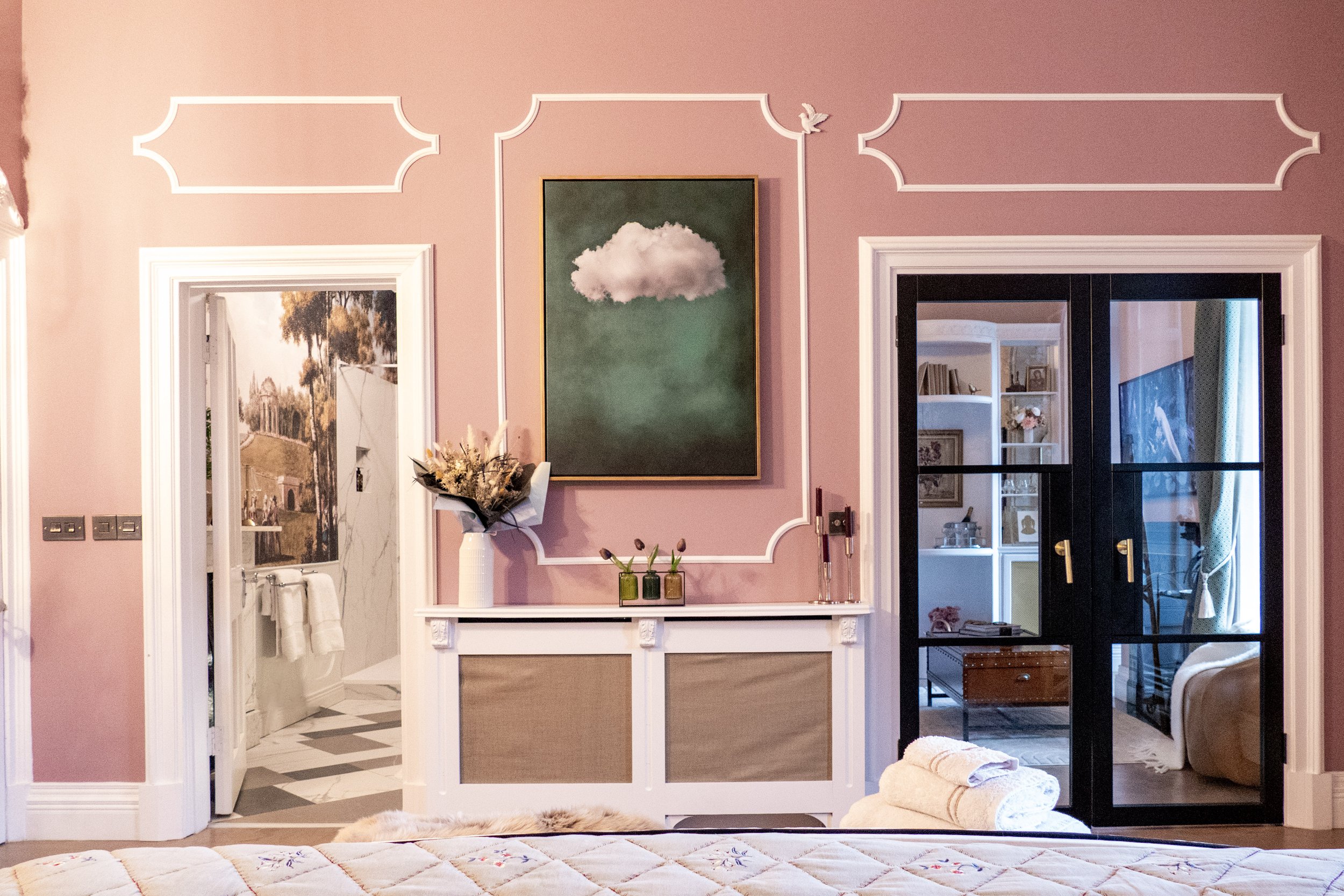 A decorated bedroom wall painted pink with white trim, featuring a large painting of a cloud, a flower arrangement, and decorative molding. Two doorways lead to a bathroom on the left and another room with a bookshelf and blue curtains on the right. 