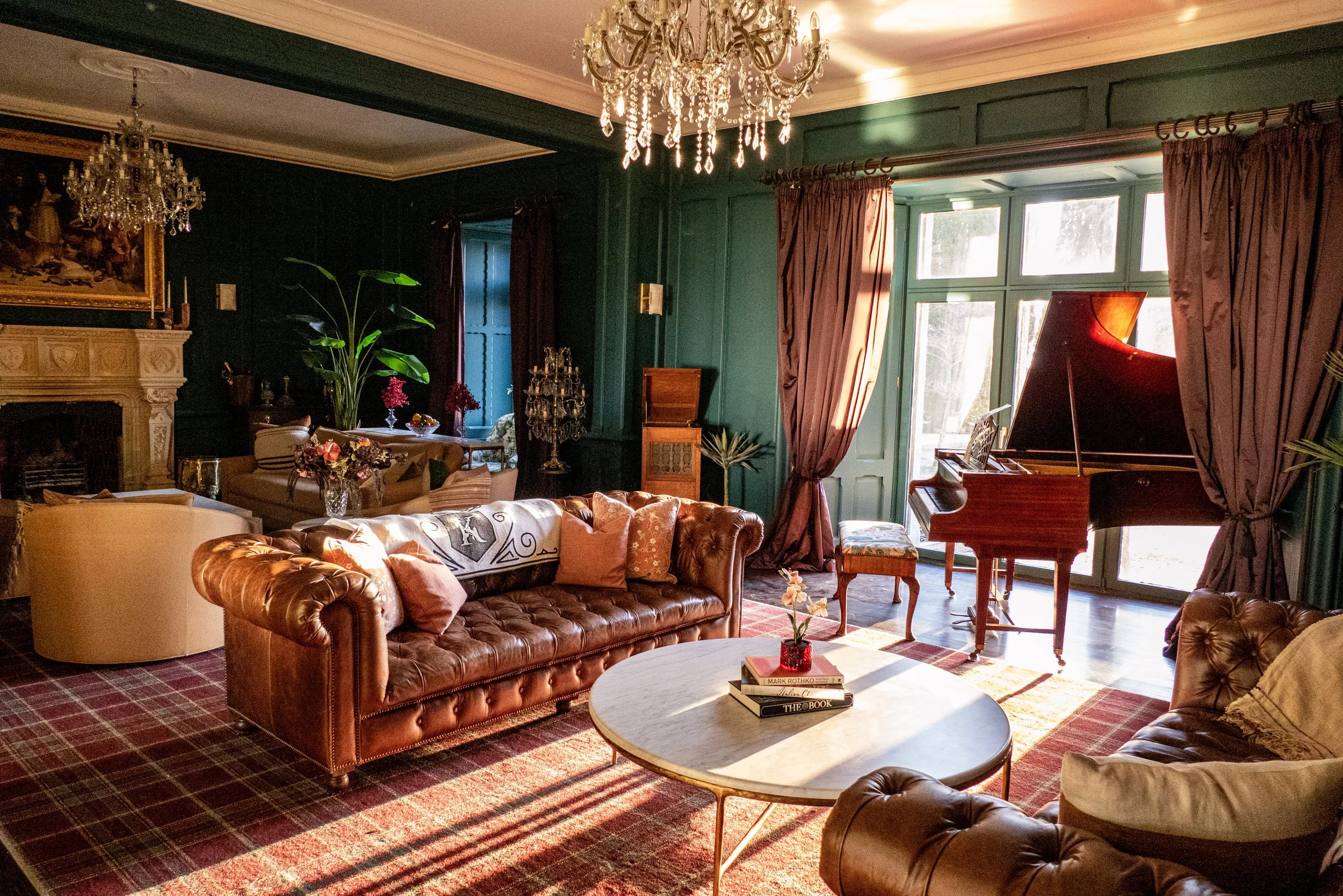 A cozy living room with green walls, a chandelier, a fireplace, a grand piano, leather sofas, a round coffee table, curtains, and various decorative items.