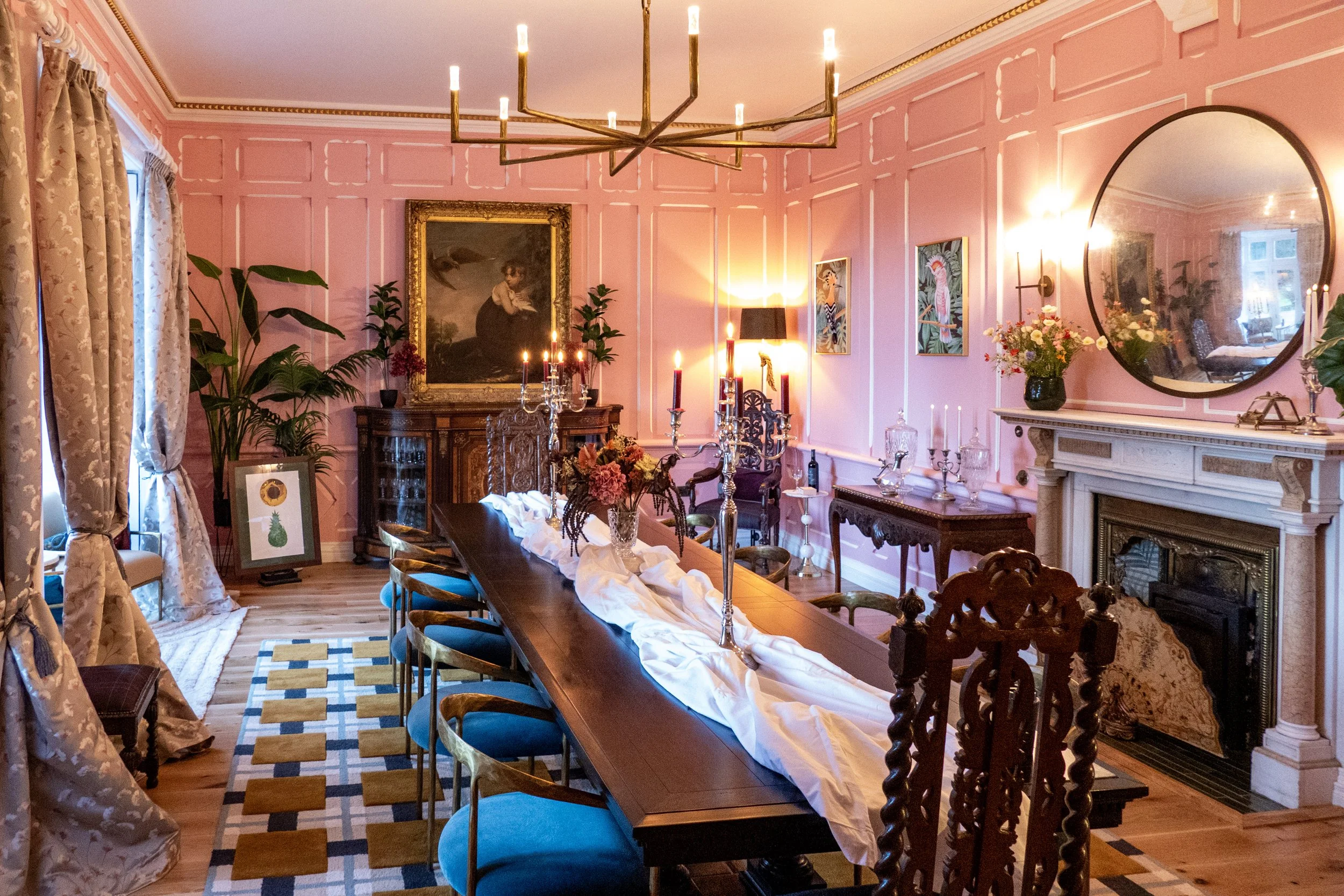 Elegant dining room with pink paneled walls, a large wooden table with chairs, floral arrangements, candelabras, a fireplace, and artwork, illuminated by warm lighting and a large chandelier.