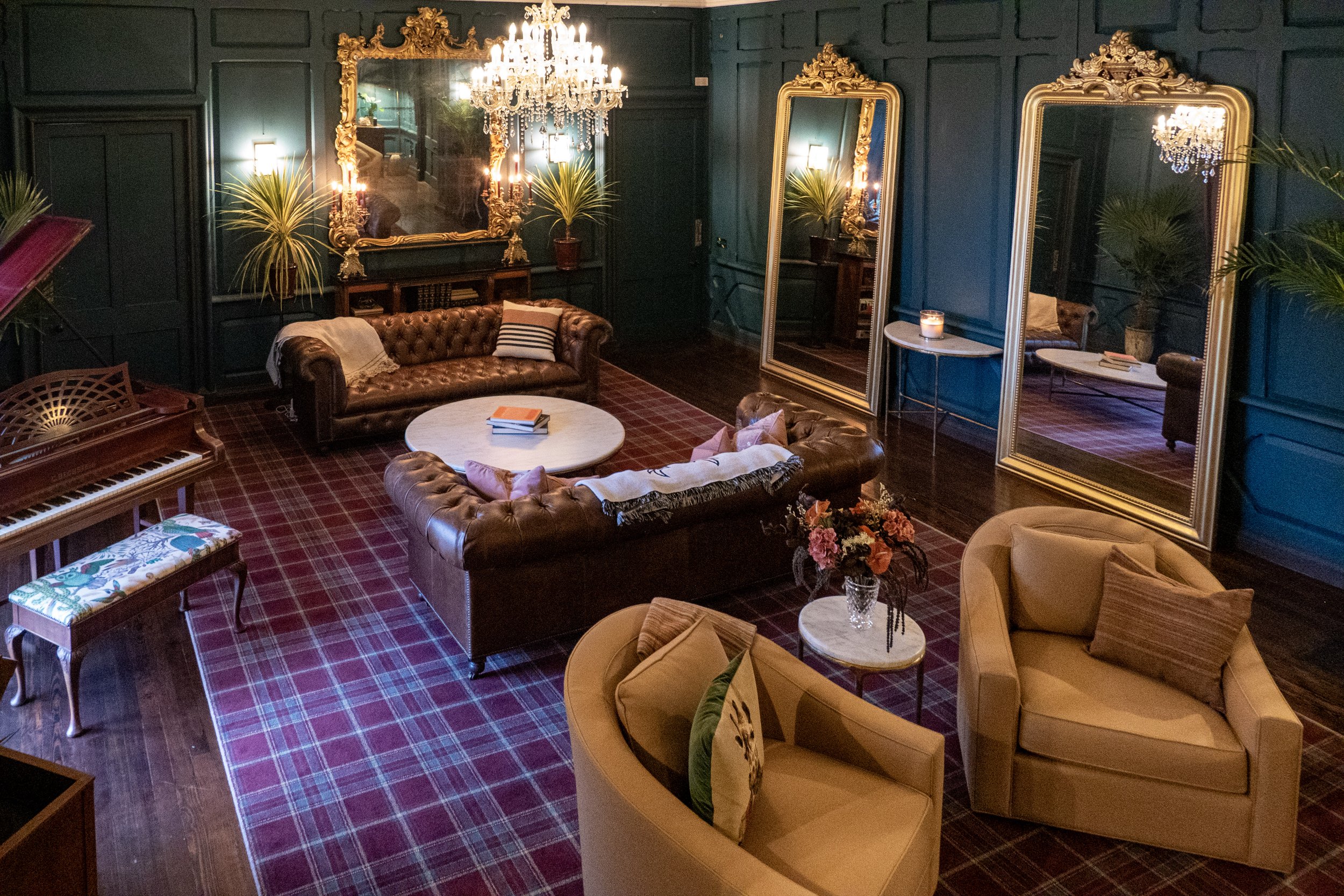 A stylish living room with dark teal paneled walls, two large ornate mirrors, a chandelier, a leather sofa, two yellow armchairs, a marble coffee table, a pink floral arrangement, a potted plant, and a grand piano with a floral cushion, all illuminat