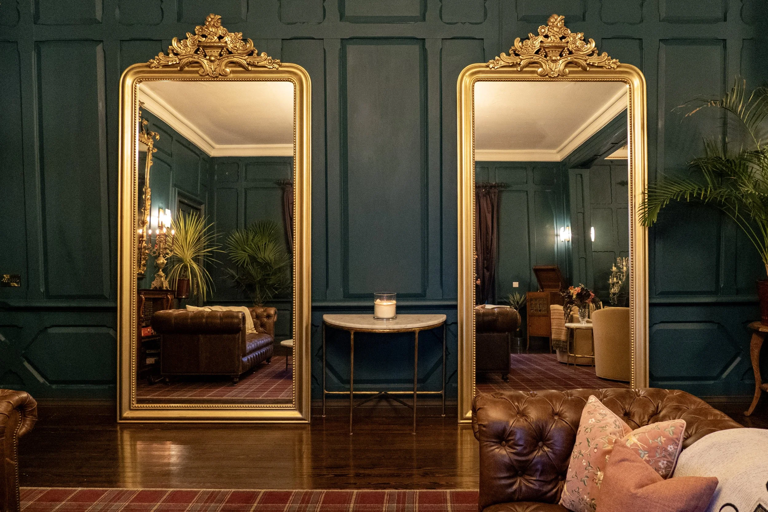 Two large ornate gold-framed mirrors are positioned against a green paneled wall, reflecting a luxurious room with leather furniture, plants, and decorative objects.