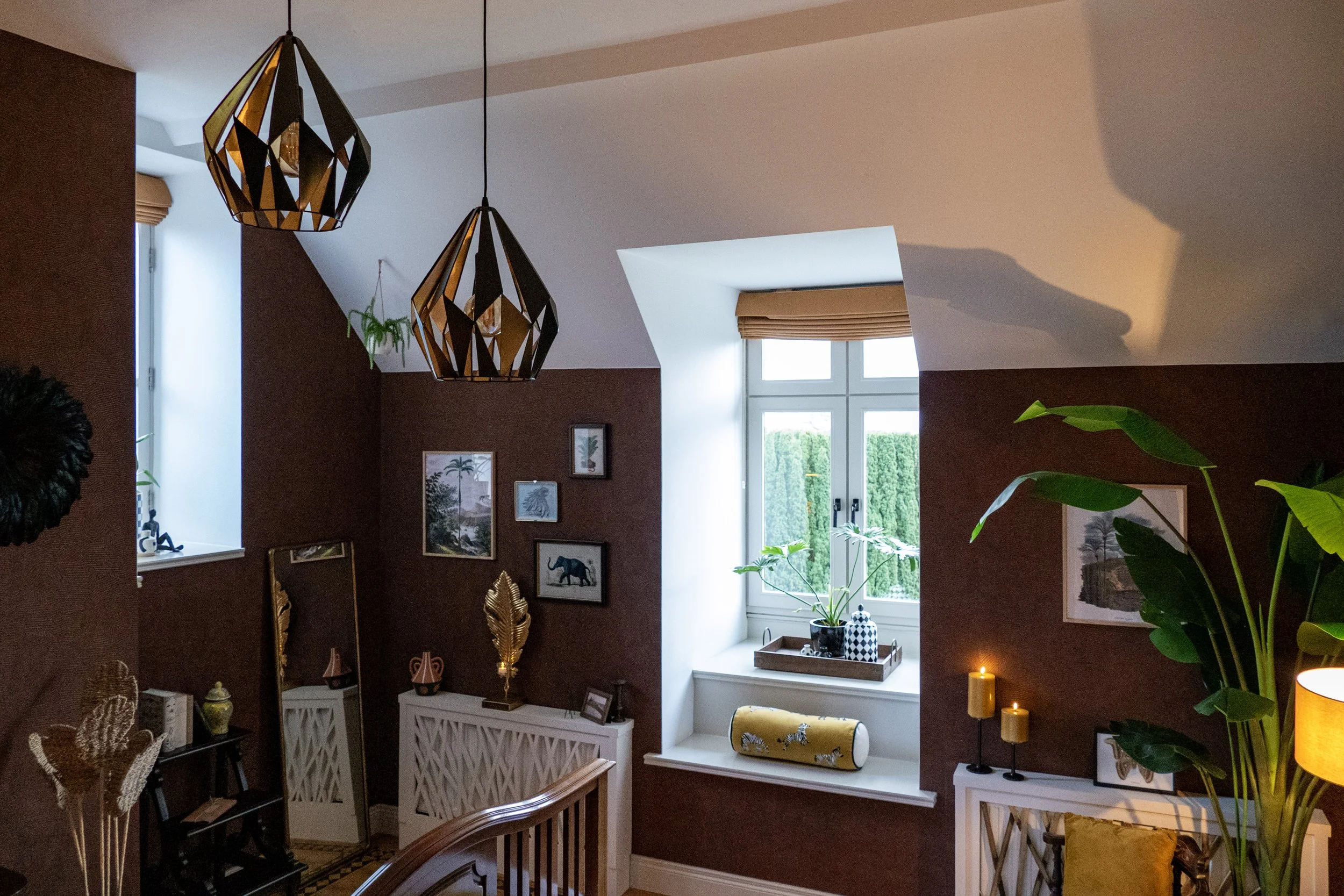 Interior of a bedroom with a white vaulted ceiling, brown walls, two black geometric pendant lights, a window with yellow blinds, green plants, framed artwork, decorative objects, candles, and yellow accents.