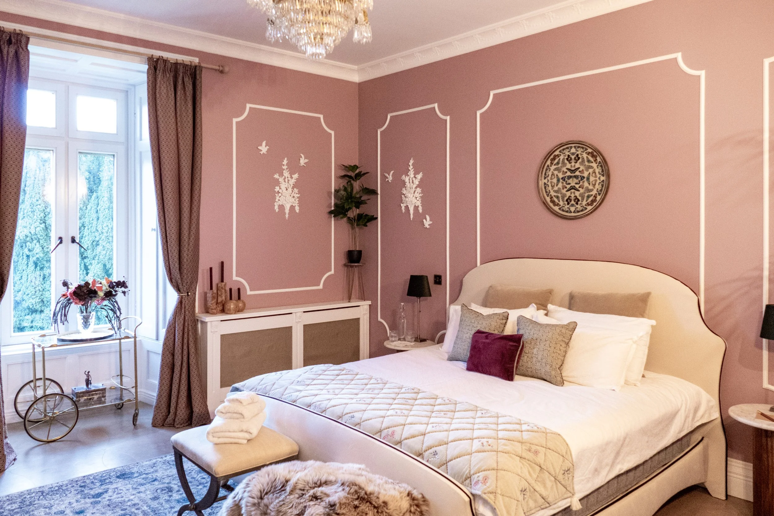 A cozy bedroom with pink walls decorated with white wall art and trim, a chandelier, a large bed with beige headboard, multiple pillows, and a cream-colored blanket. There are black table lamps on side tables, a window with brown curtains, a small ta