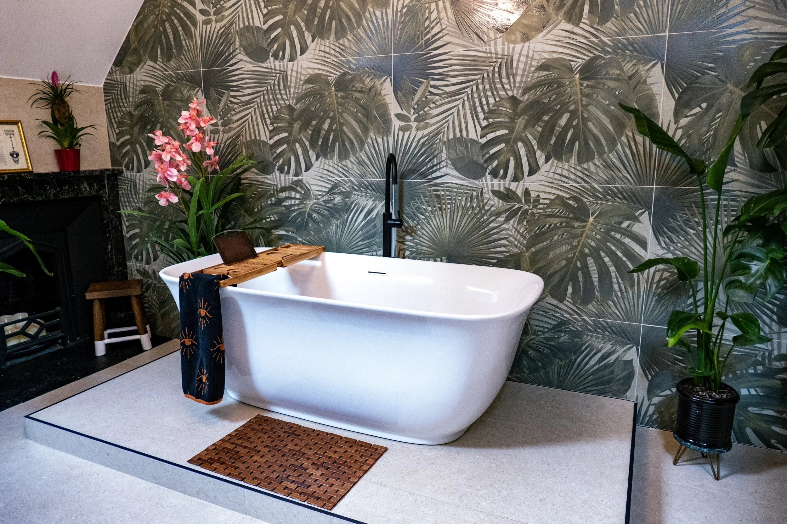 A modern bathroom features a white freestanding bathtub with a black faucet, set against a tropical leaf-patterned wall. Adjacent to the tub is a small wooden stool with a towel hanging over it, and a wooden bath caddy holding a tablet. Decor include