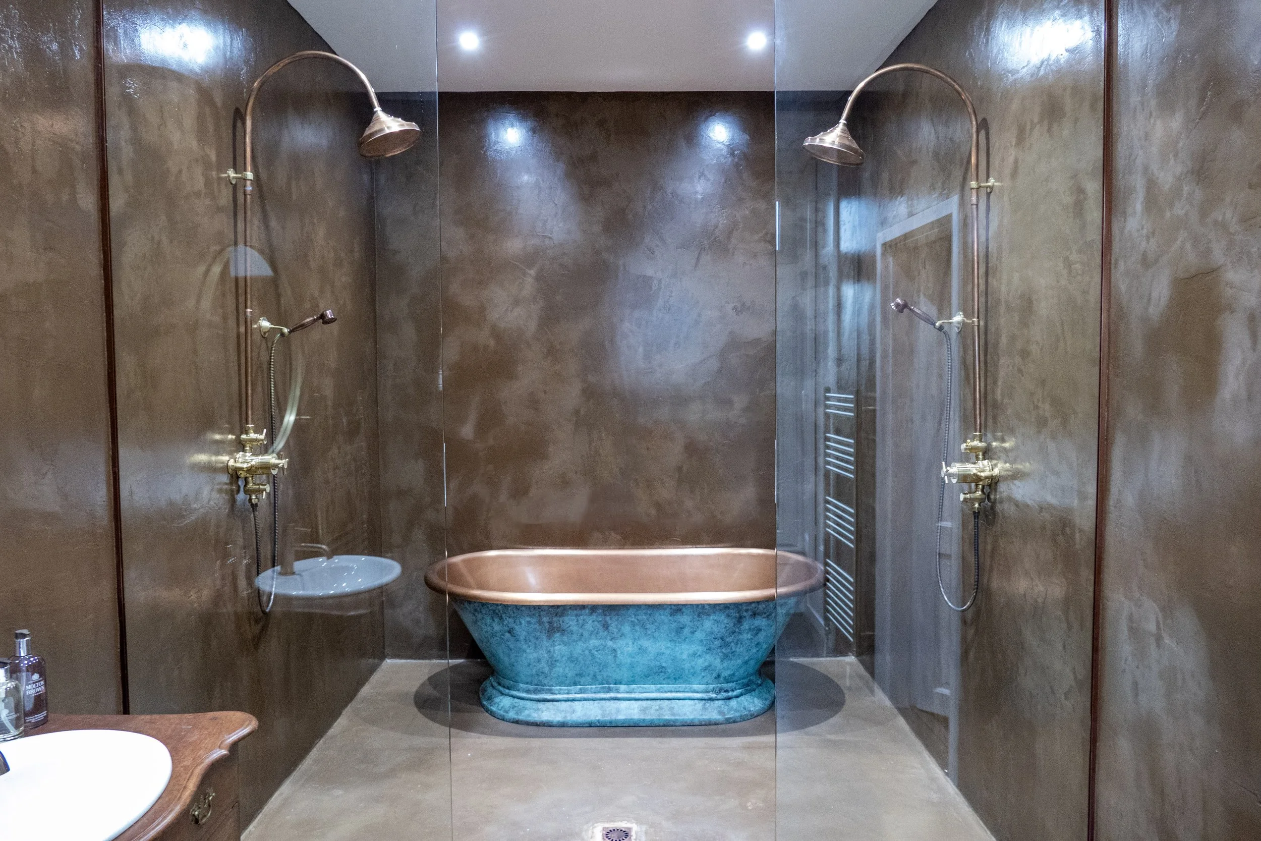 Bathroom with dark brown walls, a large freestanding bathtub with a blue base, and two separate showerheads on the walls.