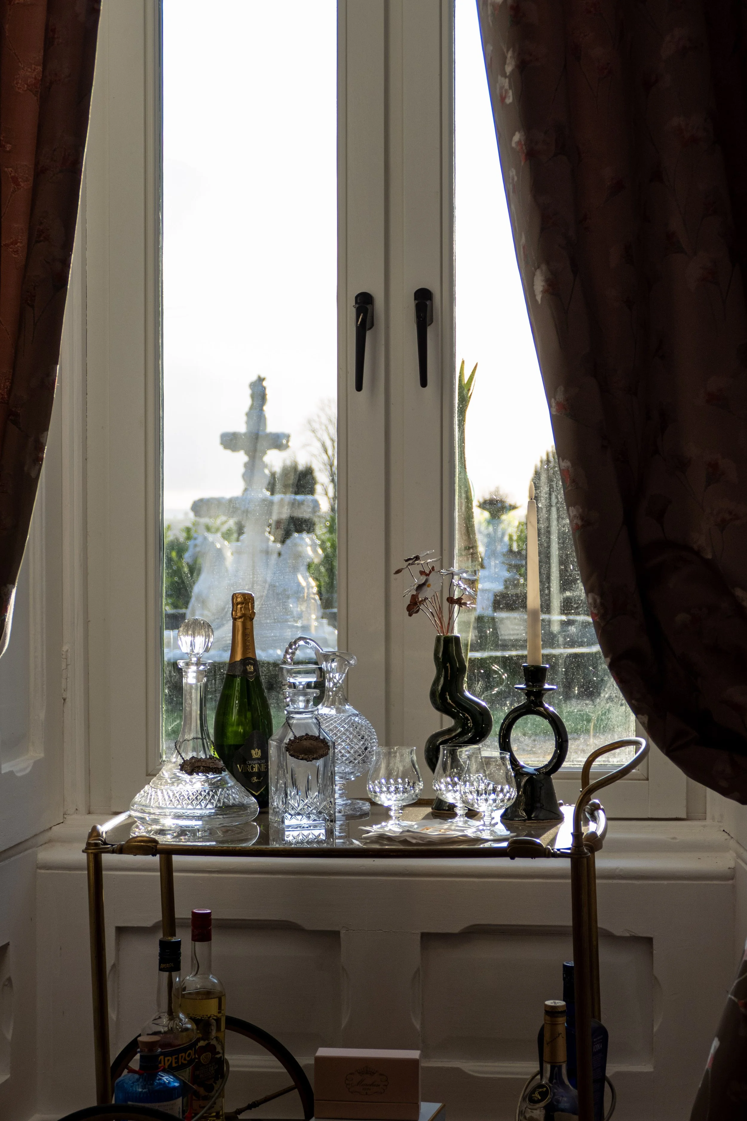 A window with curtains partially pulled aside, revealing a view of a fountain outside. On the windowsill, there are several decorative glass bottles and vases, and a tall candlestick with a candle. A bar cart with bottles of alcohol and a box is part