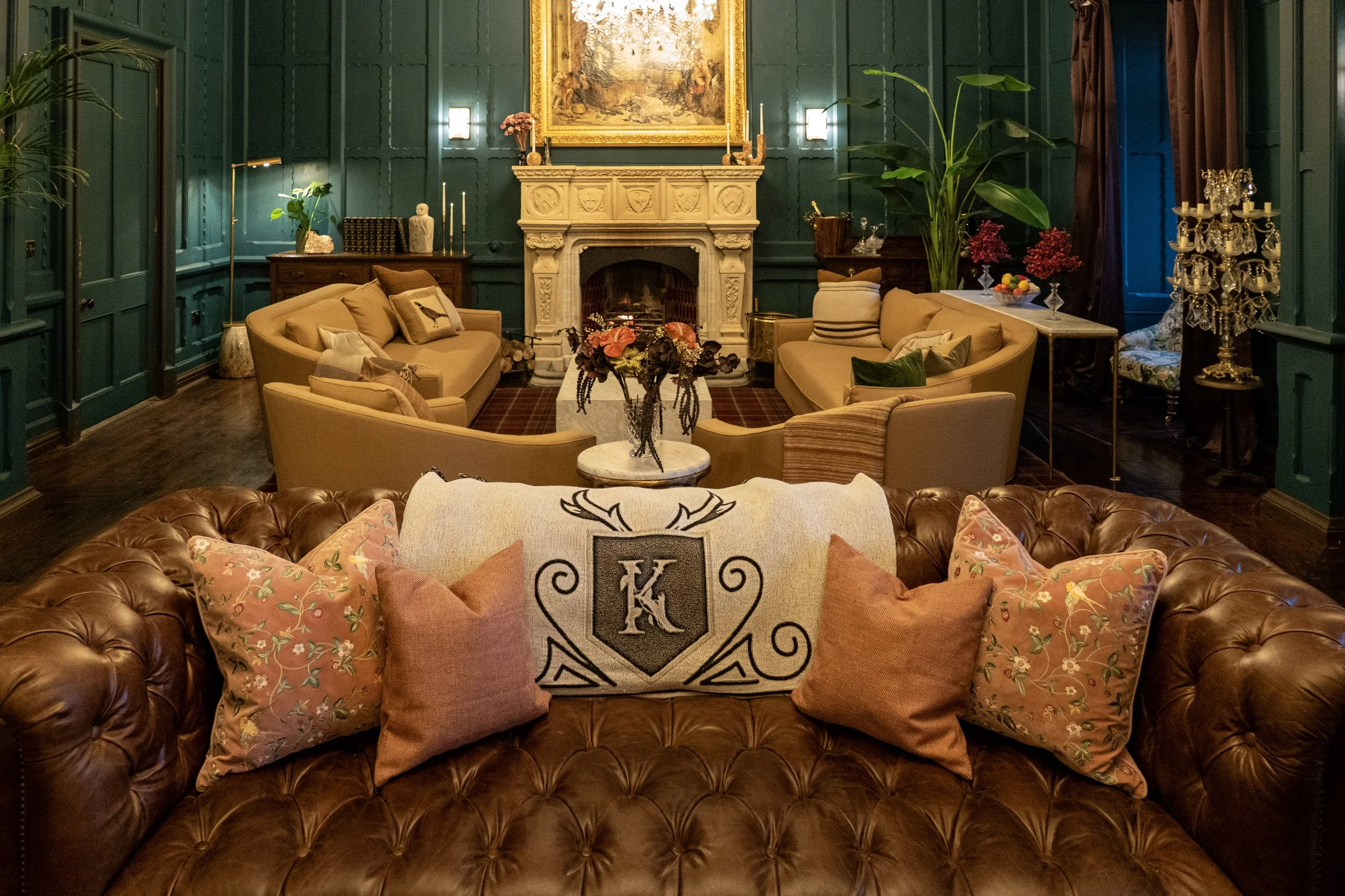 Elegant living room with dark green paneled walls, a white ornate fireplace, and a large painting above it. The room has beige and brown furniture, including a tufted leather sofa in the foreground with decorative pillows, and two curved sofas facing