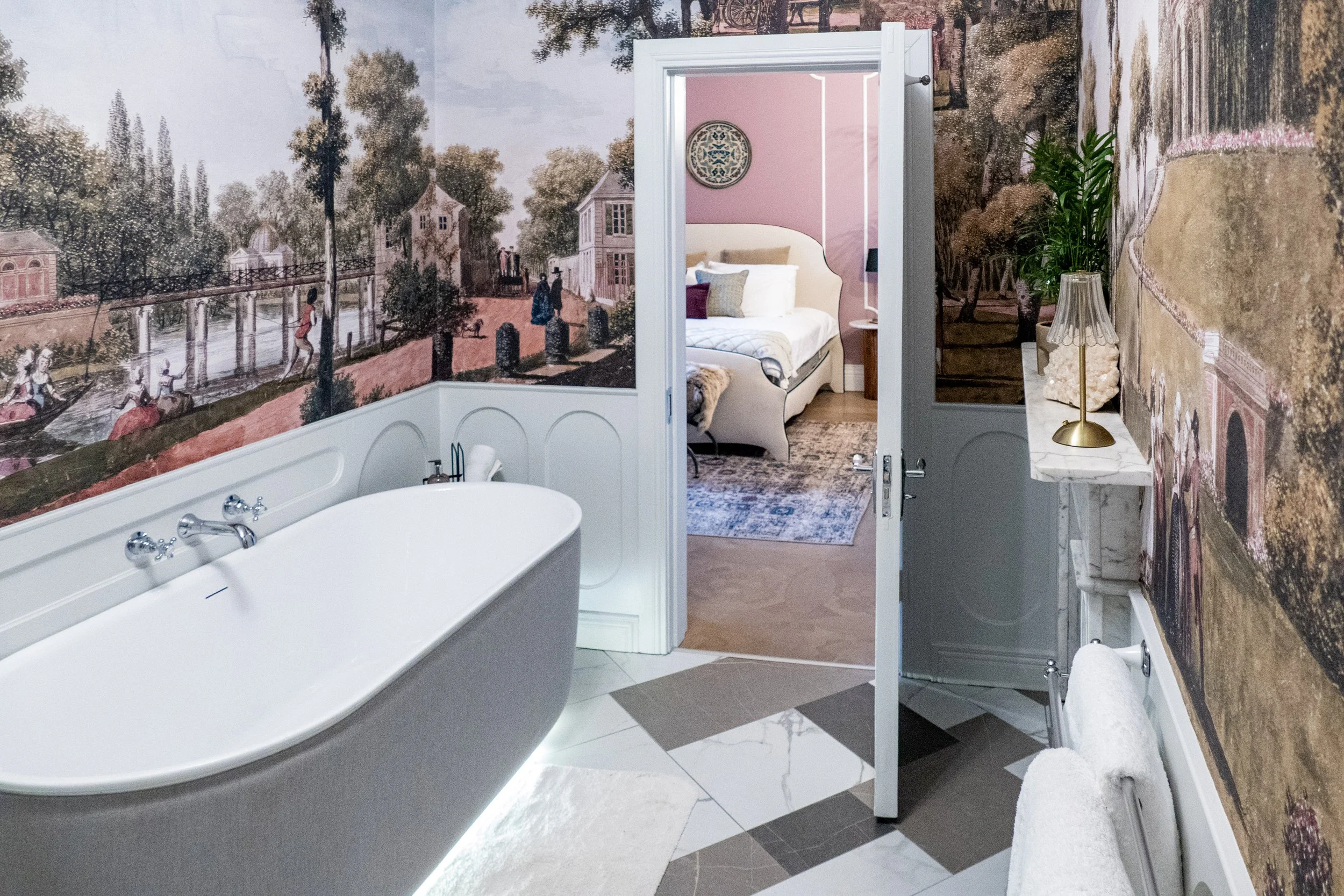 Room with large mural of a park scene and a clawfoot bathtub, leading to a bedroom with a pink wall and a bed with white bedding.