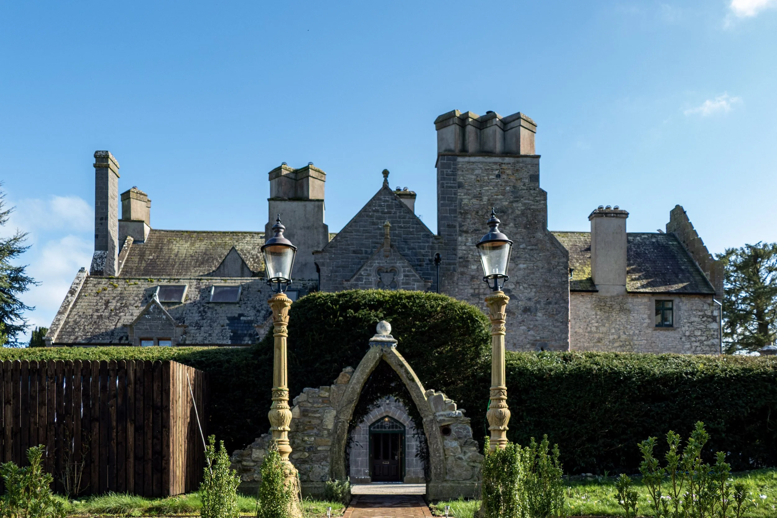 A stone castle with multiple chimneys, surrounded by a garden with two ornate street lamps and a small arched entrance in front.