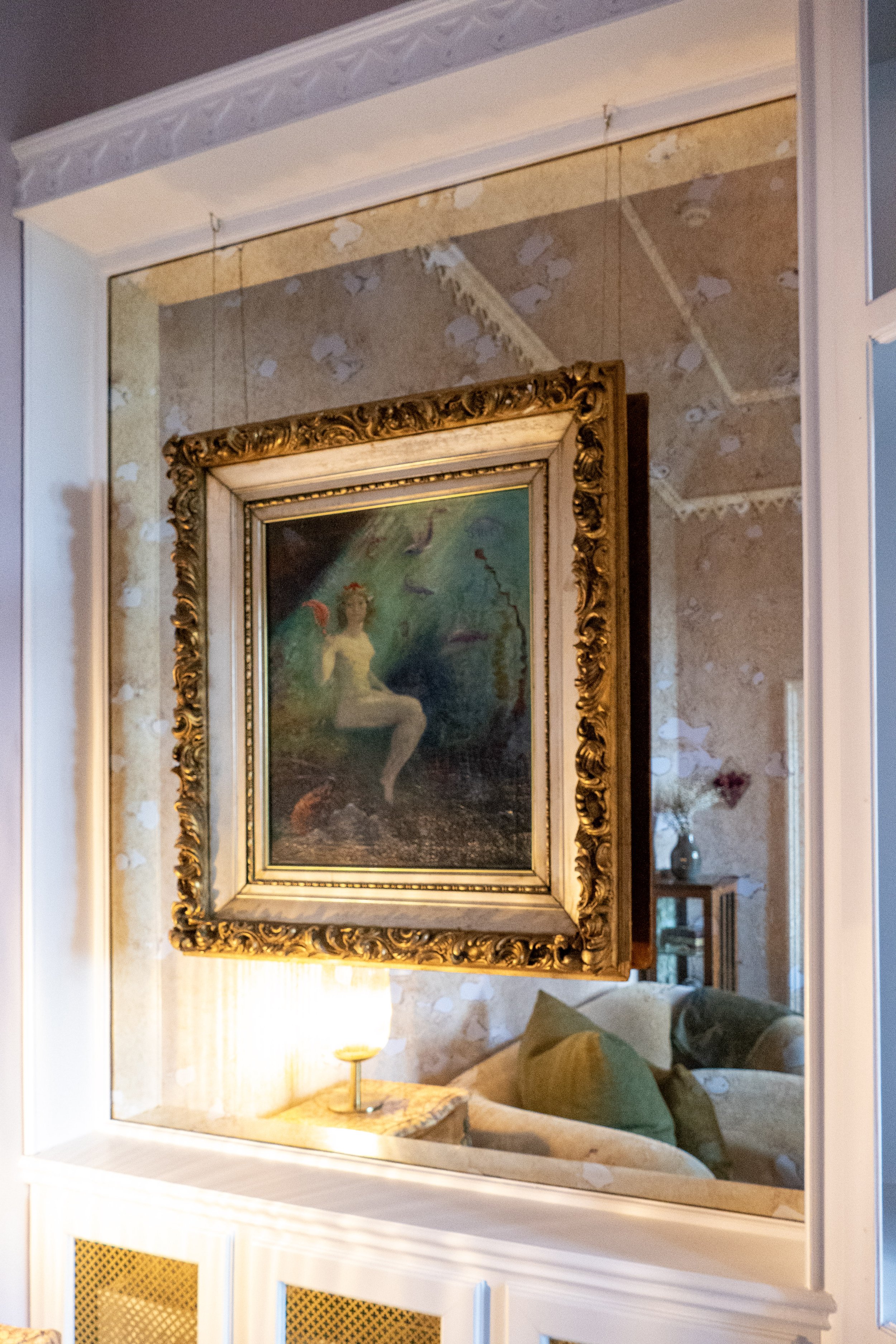 A painting of a woman sitting underwater, surrounded by fish, in a decorative gold frame, reflected in a mirror in a cozy living room.