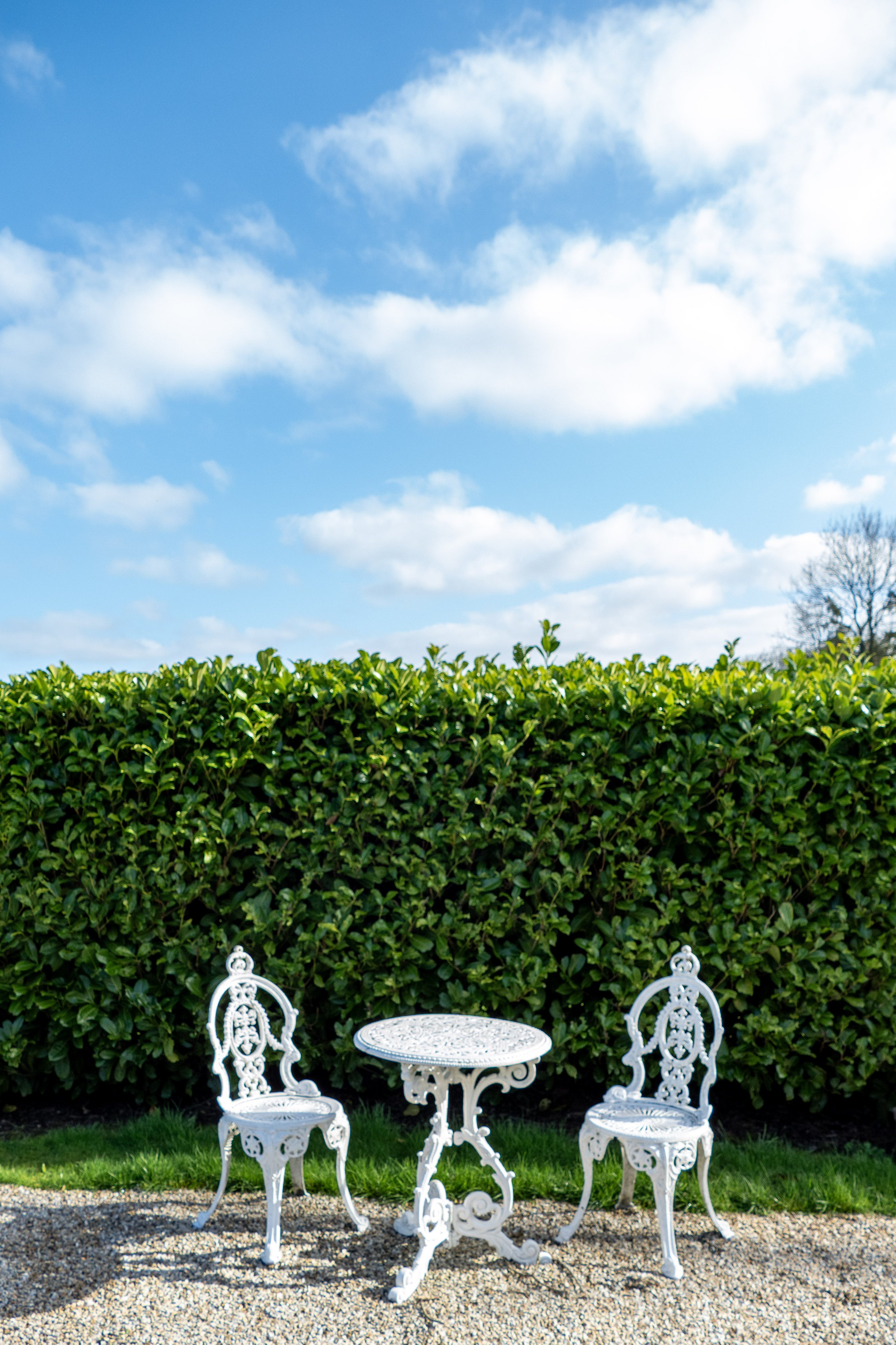 White ornate metal garden table with two matching chairs on gravel, green hedge in background, blue sky with clouds.